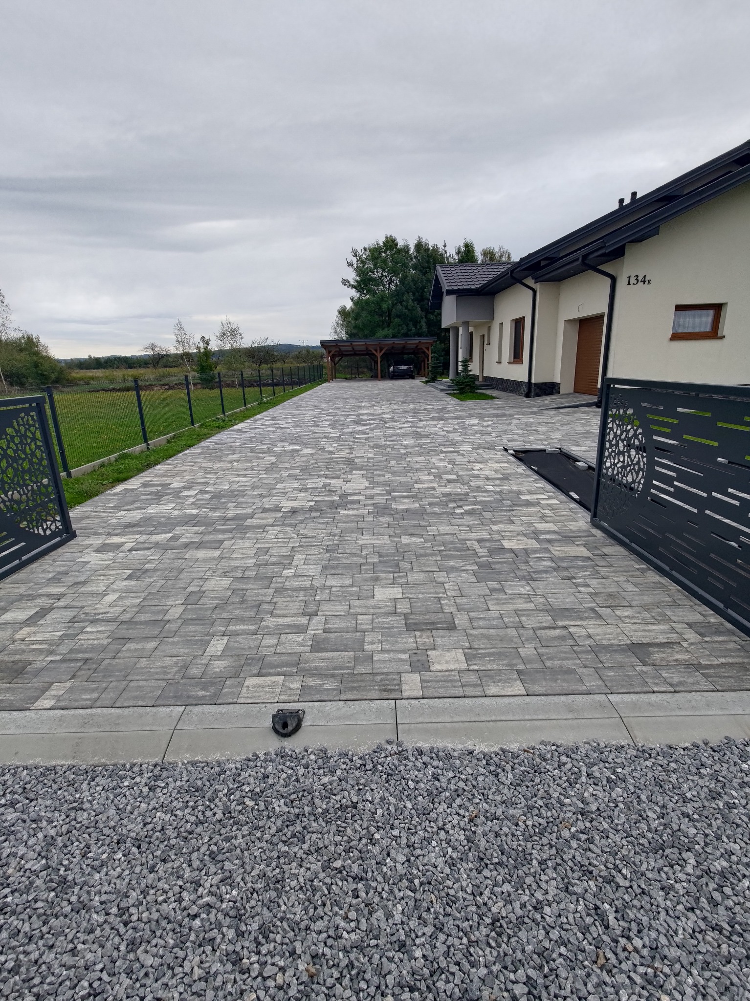 Szeroki podjazd z szarej kostki brukowej prowadzący do domu z numerem 134a, widok zza otwartej, ozdobnej bramy, po bokach trawnik i ogrodzenie, w tle zieleń i pochmurne niebo.