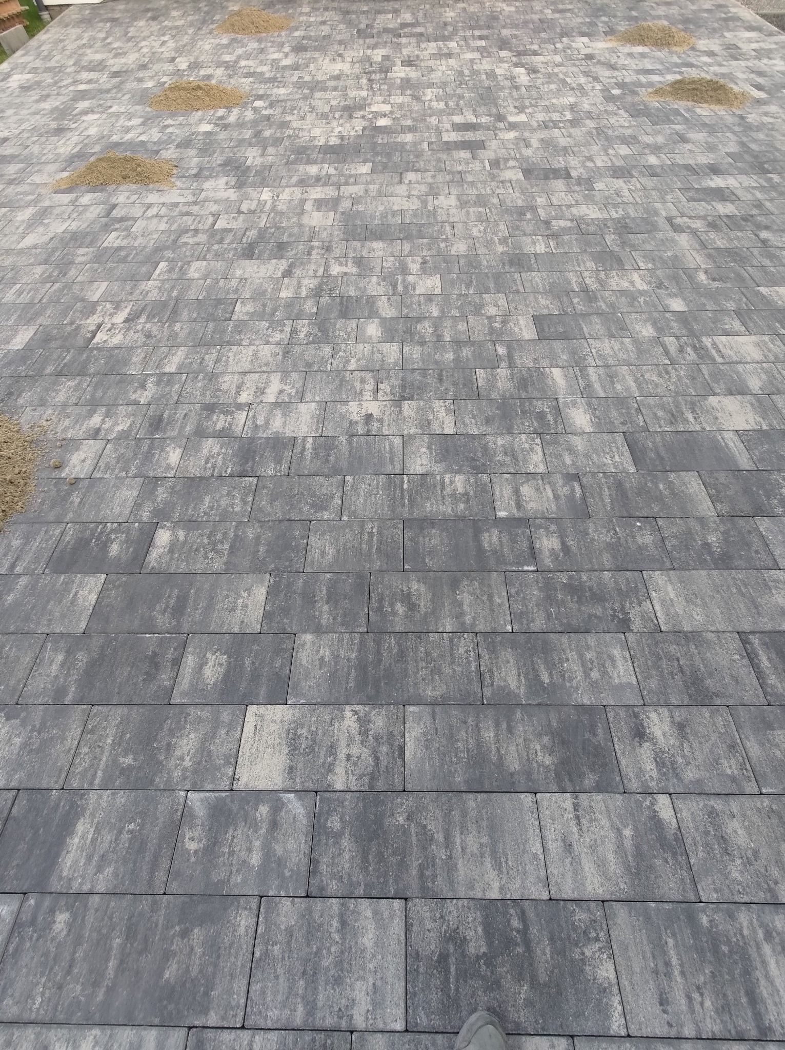 Nowo ułożona, szara kostka brukowa o prostokątnym kształcie z widocznymi trzema małymi pryzmami piasku do wypełnienia fug. Jeden but w lewym dolnym rogu kadru sugeruje skalę.