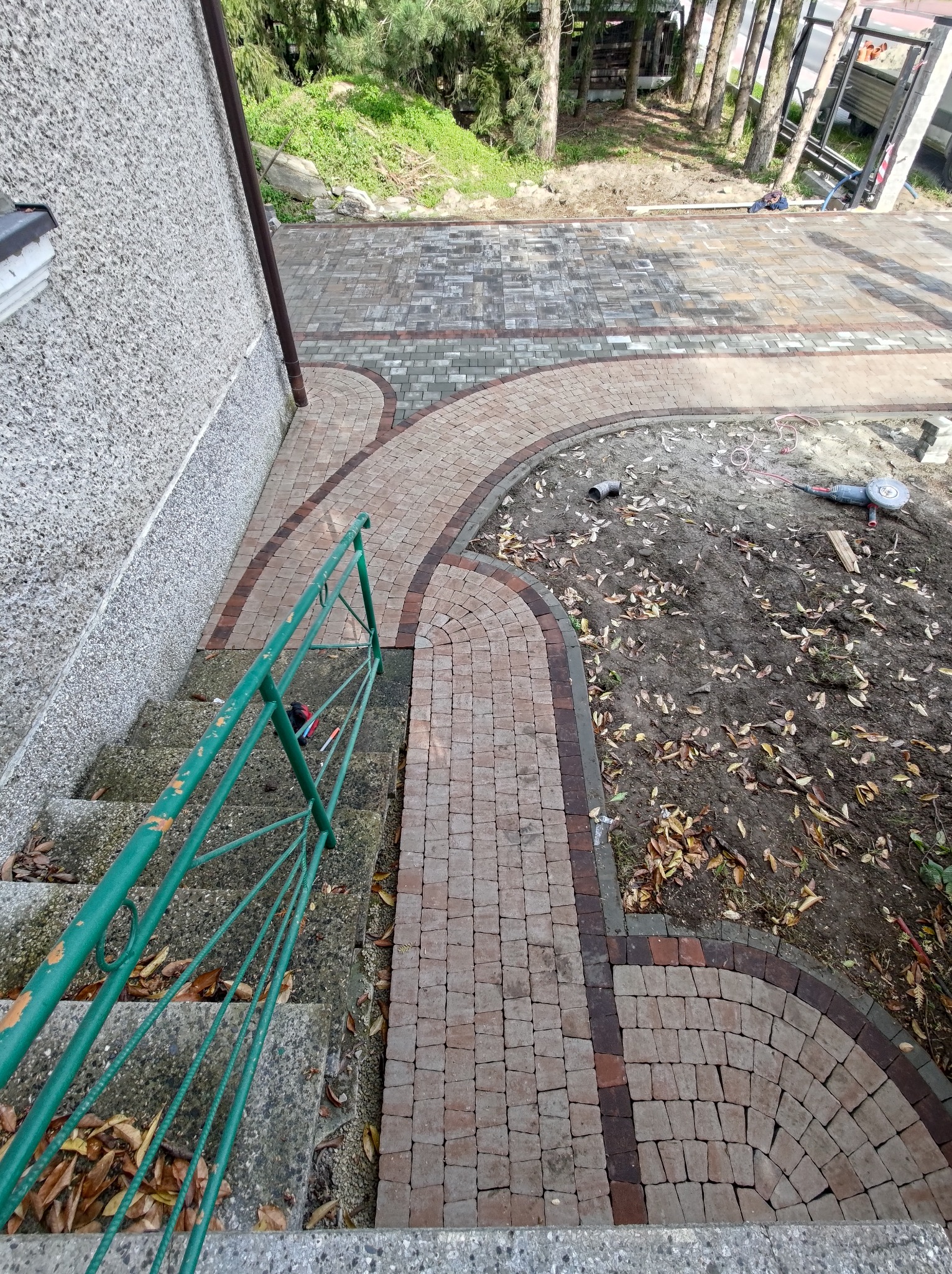 Widok z góry na fragment posesji z ułożoną kostką brukową w odcieniach szarości i brązu, tworzącą ścieżkę z zakrzywieniami, obok schodów z zieloną metalową barierką oraz obszaru ziemi z liśćmi...