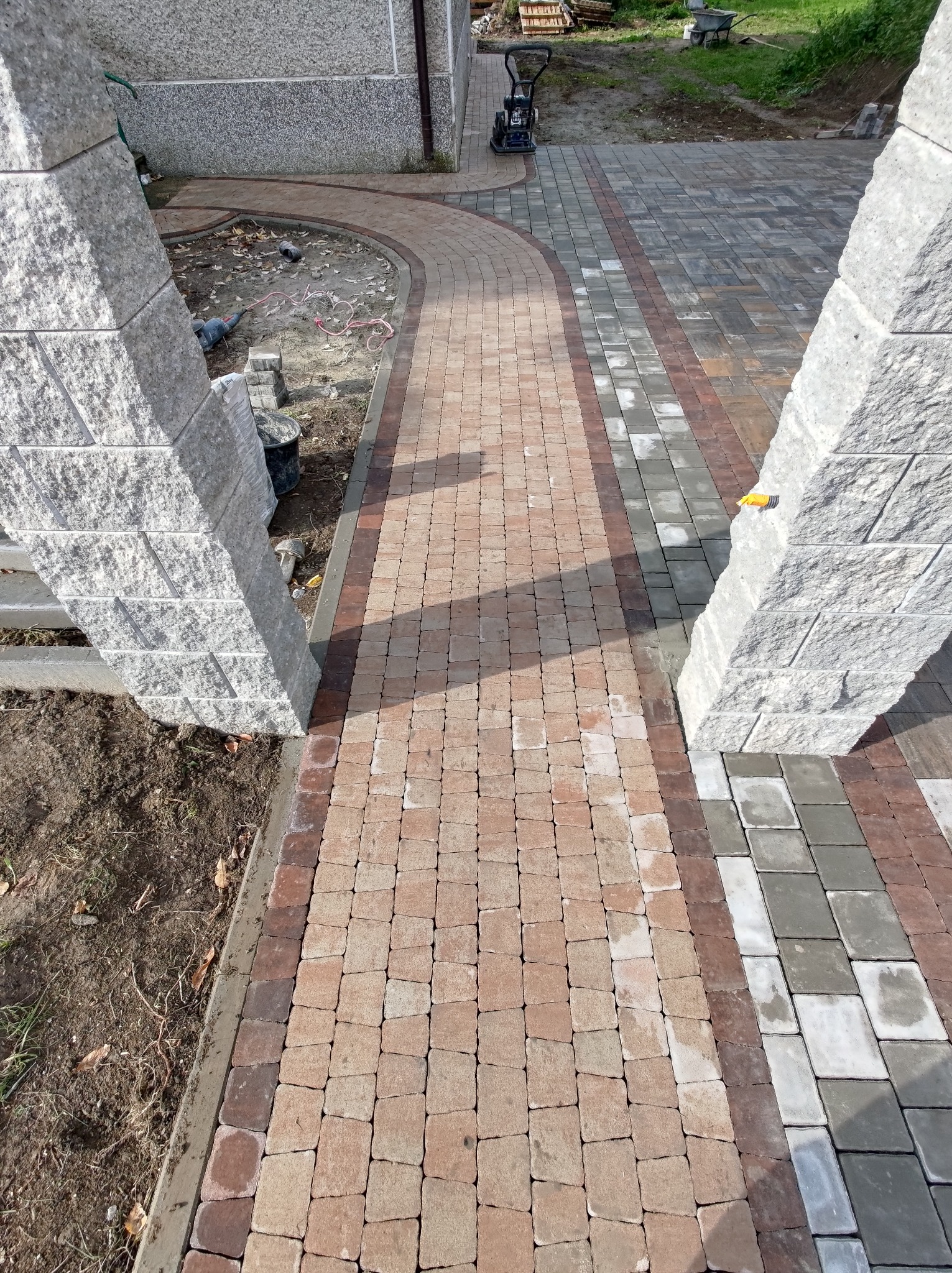 Ułożona ścieżka z kostki brukowej w trzech kolorach, prowadząca między dwoma kamiennymi filarami, z ubijarką w tle.