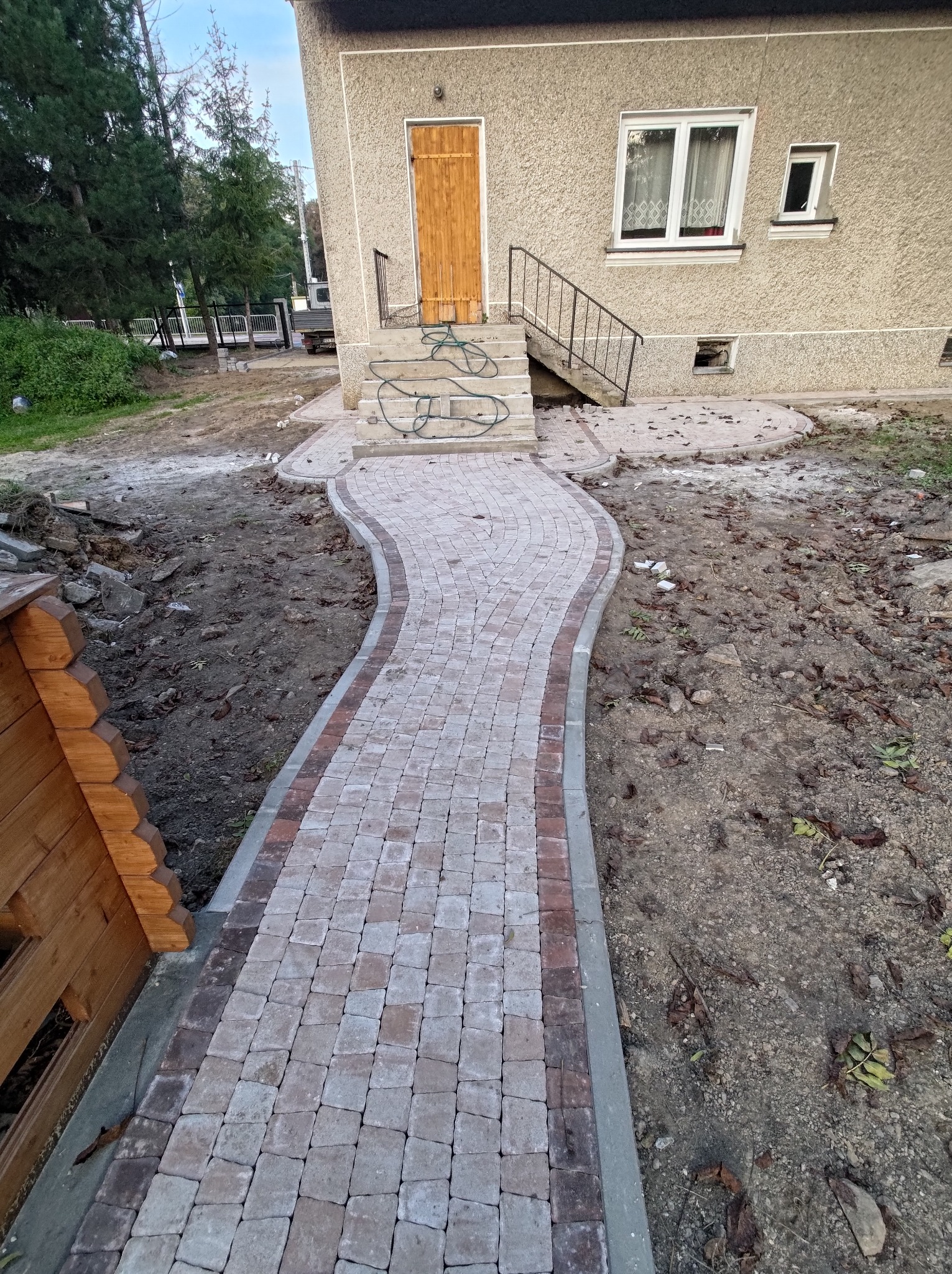 Kaskadowa ścieżka z kostki brukowej prowadząca do wejścia domu, z widocznymi elementami krajobrazu w trakcie prac wykończeniowych.