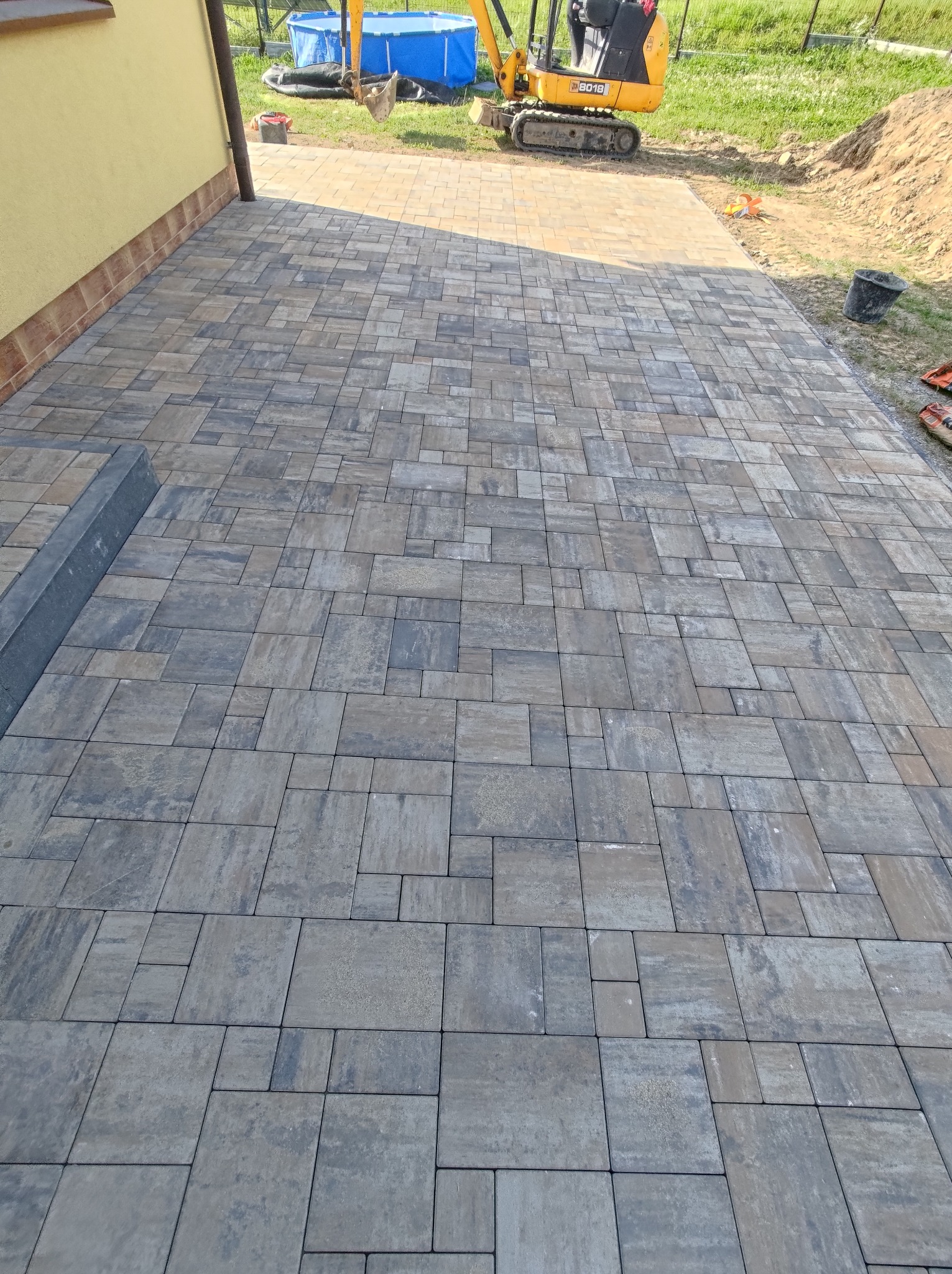 Ułożona kostka brukowa w odcieniach szarości i brązu, z widocznym fragmentem budynku i mini koparką w tle.