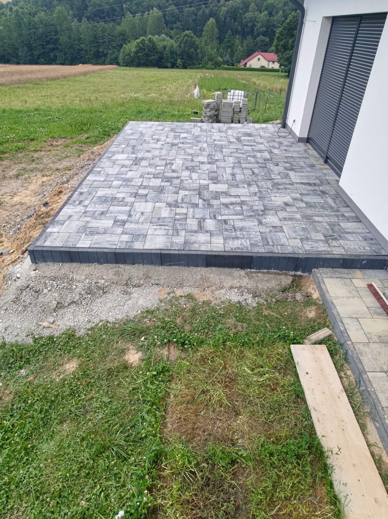 Nowo ułożony, szary taras z kostki brukowej z ciemnoszarą obwódką, przylegający do białego budynku z szarymi roletami, widok na zielone pole i las w tle, prace w toku.