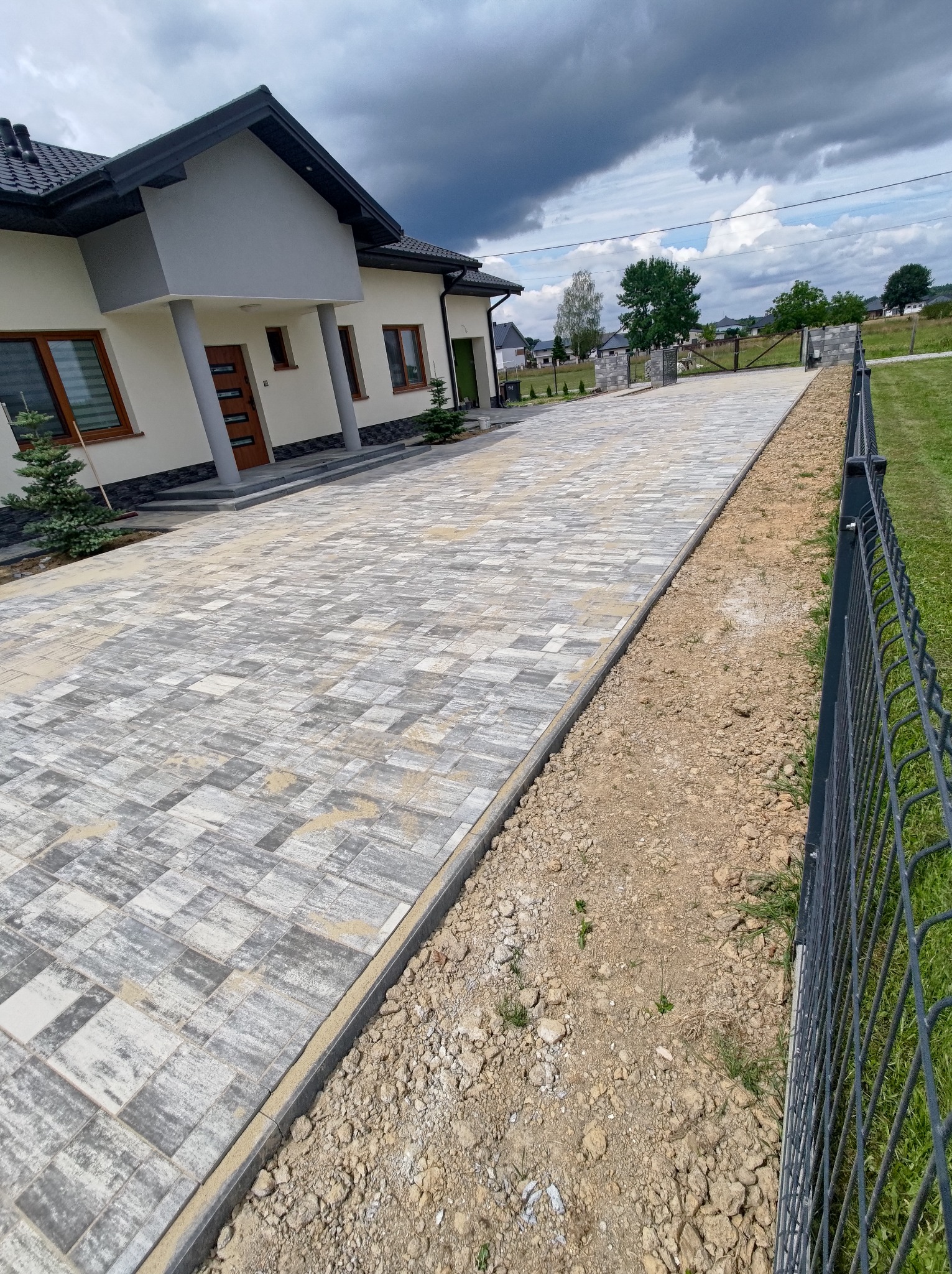 Nowoczesny podjazd z kostki brukowej w odcieniach szarości i beżu prowadzący do domu z beżową elewacją i ciemną stolarką okienną, widok na posesję z metalowym ogrodzeniem.