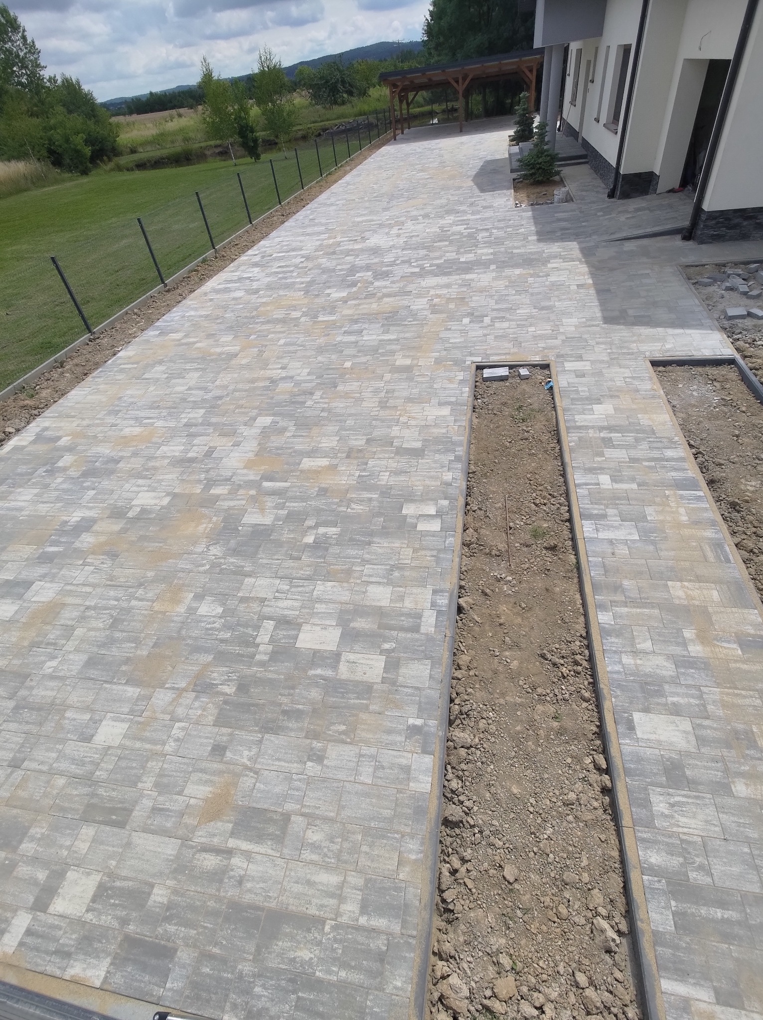 Długa, szara kostka brukowa ułożona przed domem, z widocznymi przerwami na rośliny, częściowo jeszcze wypełnionymi ziemią, w tle zielony krajobraz.