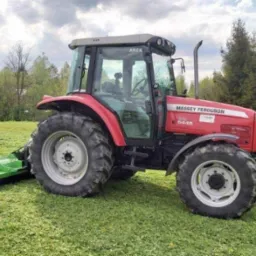 Czerwony traktor Massey Ferguson 5445 z zieloną kosiarką rotacyjną na nierównym, świeżo skoszonym trawniku, widok z boku, w tle drzewa i pochmurne niebo.