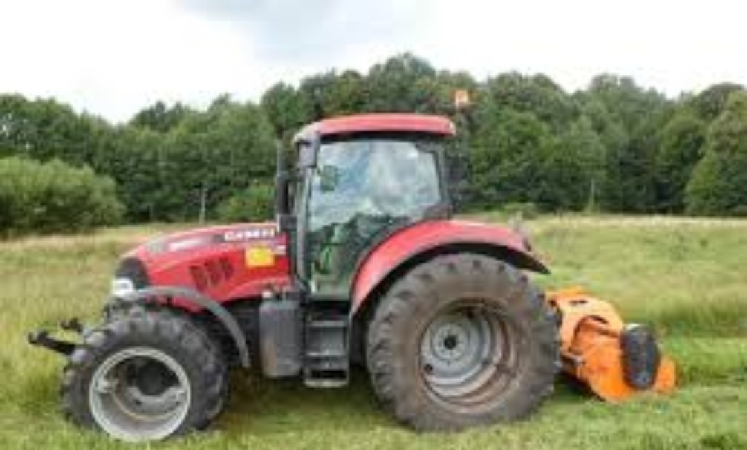 Czerwony traktor Case IH z pomarańczową kosiarką rotacyjną na zarośniętym polu z drzewami w tle.