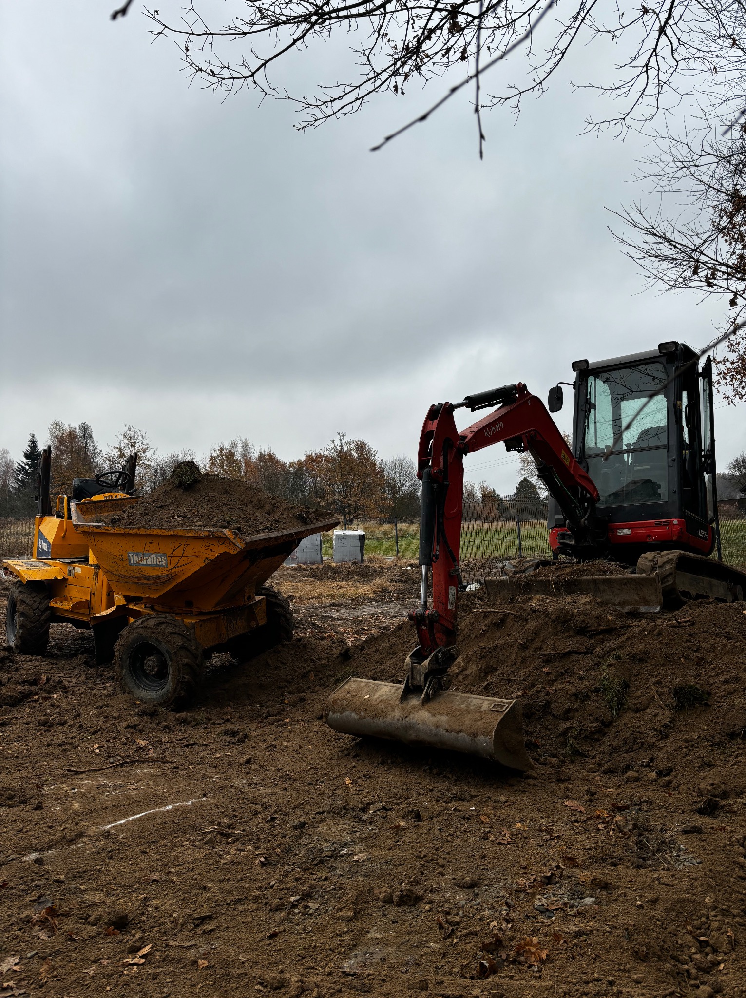 Żółta wywrotka z ziemią i czerwona koparka Kubota na placu budowy, jesienne drzewa w tle. Przygotowanie terenu pod inwestycję.
