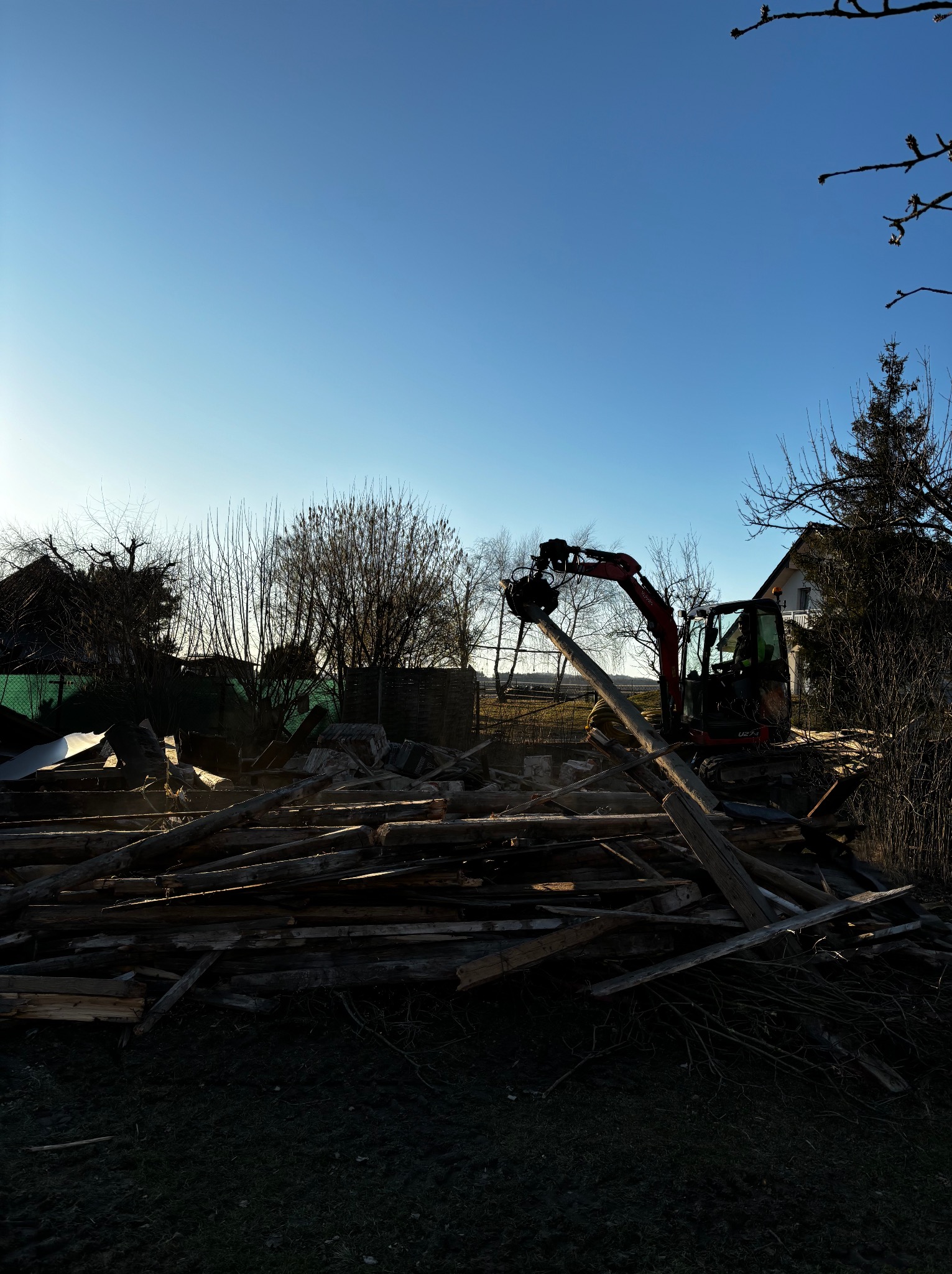 Czerwona koparka z chwytakiem podnosi drewniane belki na tle gruzowiska i błękitnego nieba. Widoczny fragment budynku w oddali. Prace rozbiórkowe w toku.