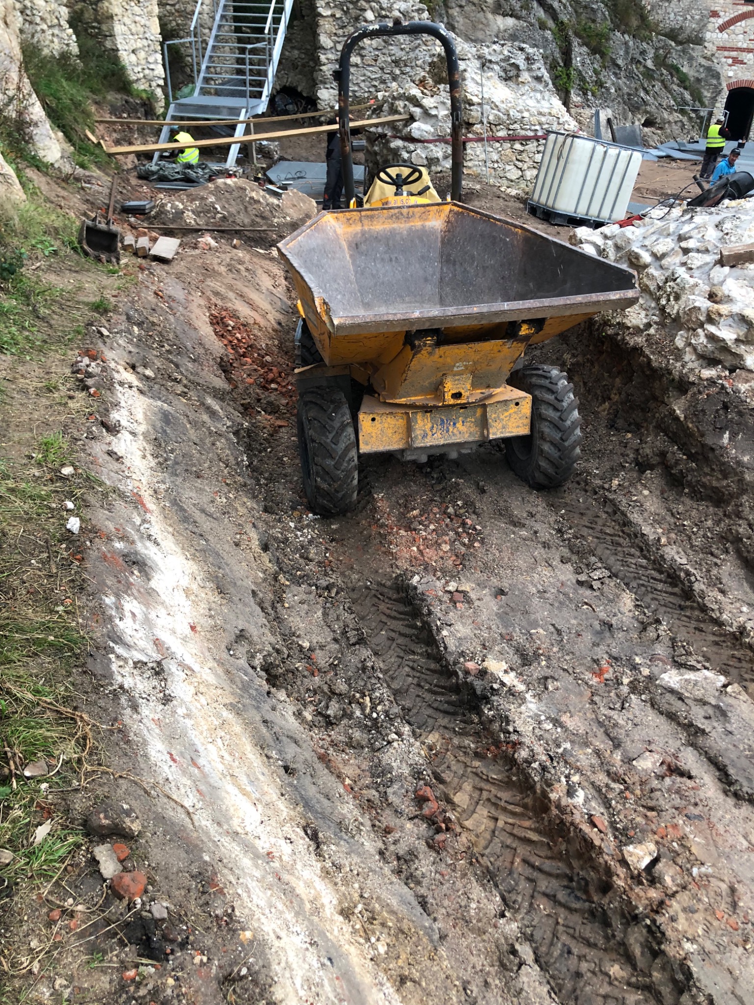 Żółta wywrotka na dwóch kołach z ładunkiem na wykopie z gruzem ceglanym, ślady opon w błocie, w tle mur z kamienia i pracownicy budowlani.