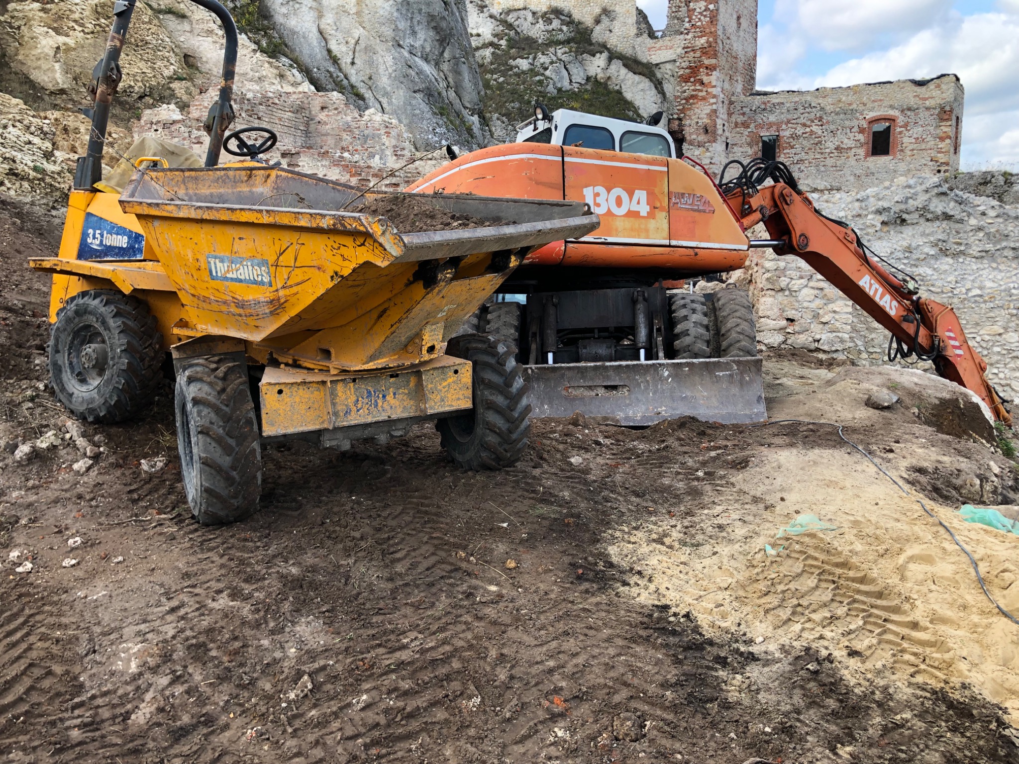 Żółta wywrotka Thwaites 3.5 tonne i pomarańczowa koparka Atlas 1304 na placu budowy z ruinami zamku w tle, ślady opon na błotnistym terenie.