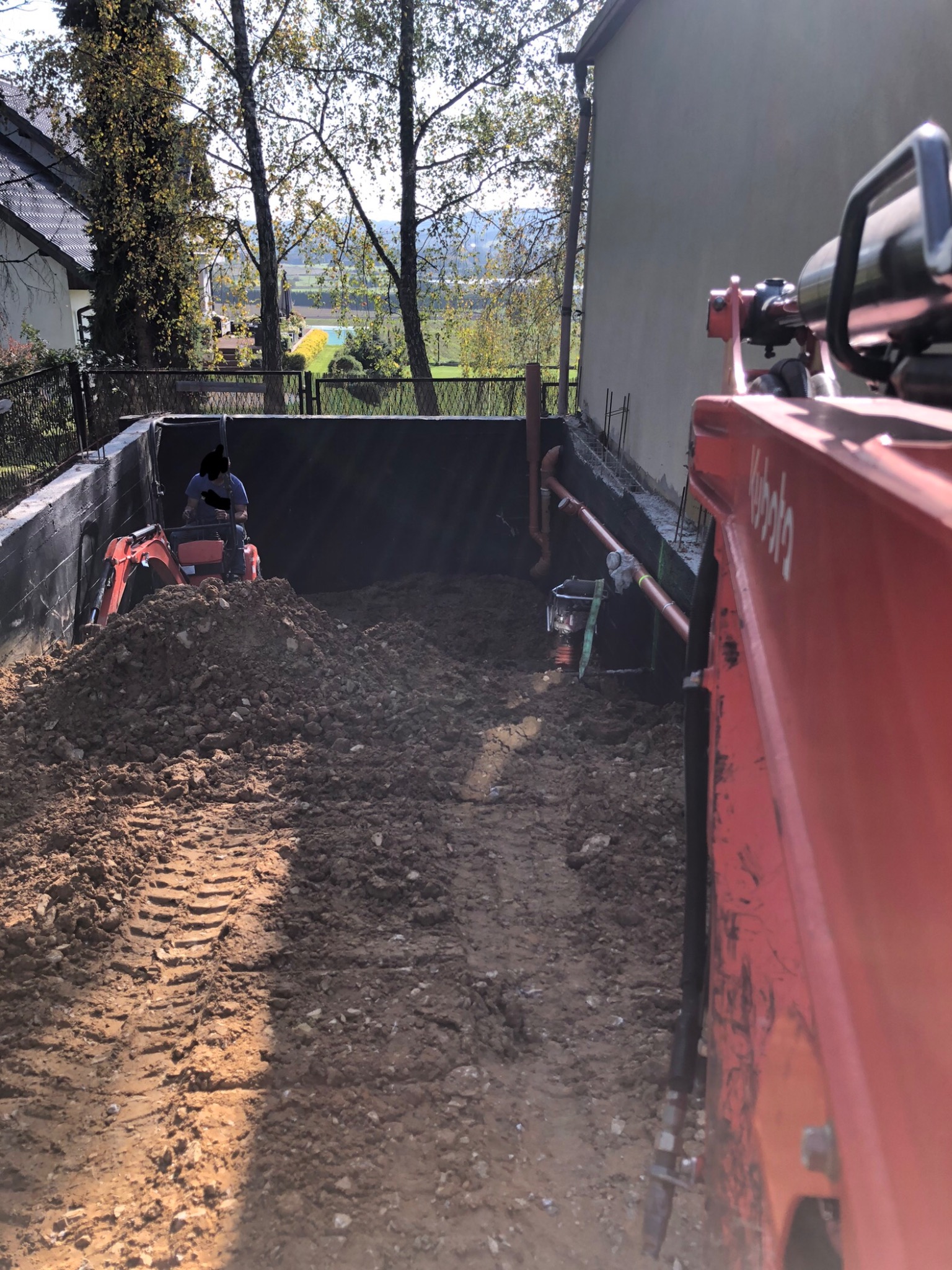 Mała koparka Kubota z operatorem pracuje w wykopie obok ściany budynku, widoczne ślady opon na ziemi i rury odprowadzające wodę w tle.