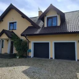 Żółty dom jednorodzinny z ciemnobrązowym dachem i dwoma garażami, widok z brukowanego podjazdu w słoneczny dzień.