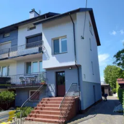 Dwukondygnacyjny dom po odświeżeniu elewacji w odcieniach szarości i bieli, z balkonami z kutymi balustradami, schodami wejściowymi z terakoty i stertą rusztowań obok, na brukowanym podjeździe...