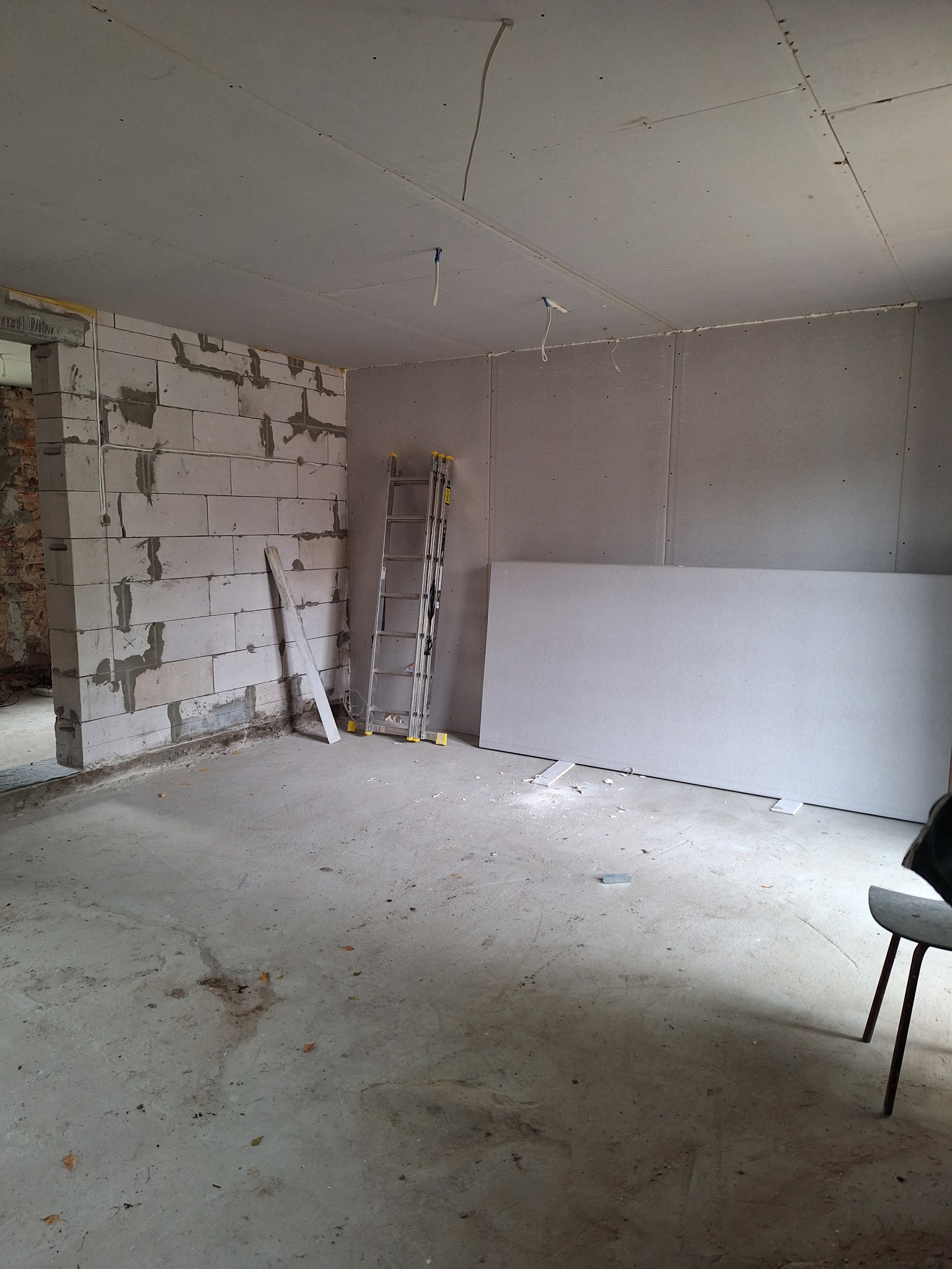 Surowe wnętrze w trakcie prac: ściany z bloków betonowych, częściowo pokryte płytami gipsowo-kartonowymi, aluminiowa drabina oparta o ścianę, widoczne przewody pod sufitem.