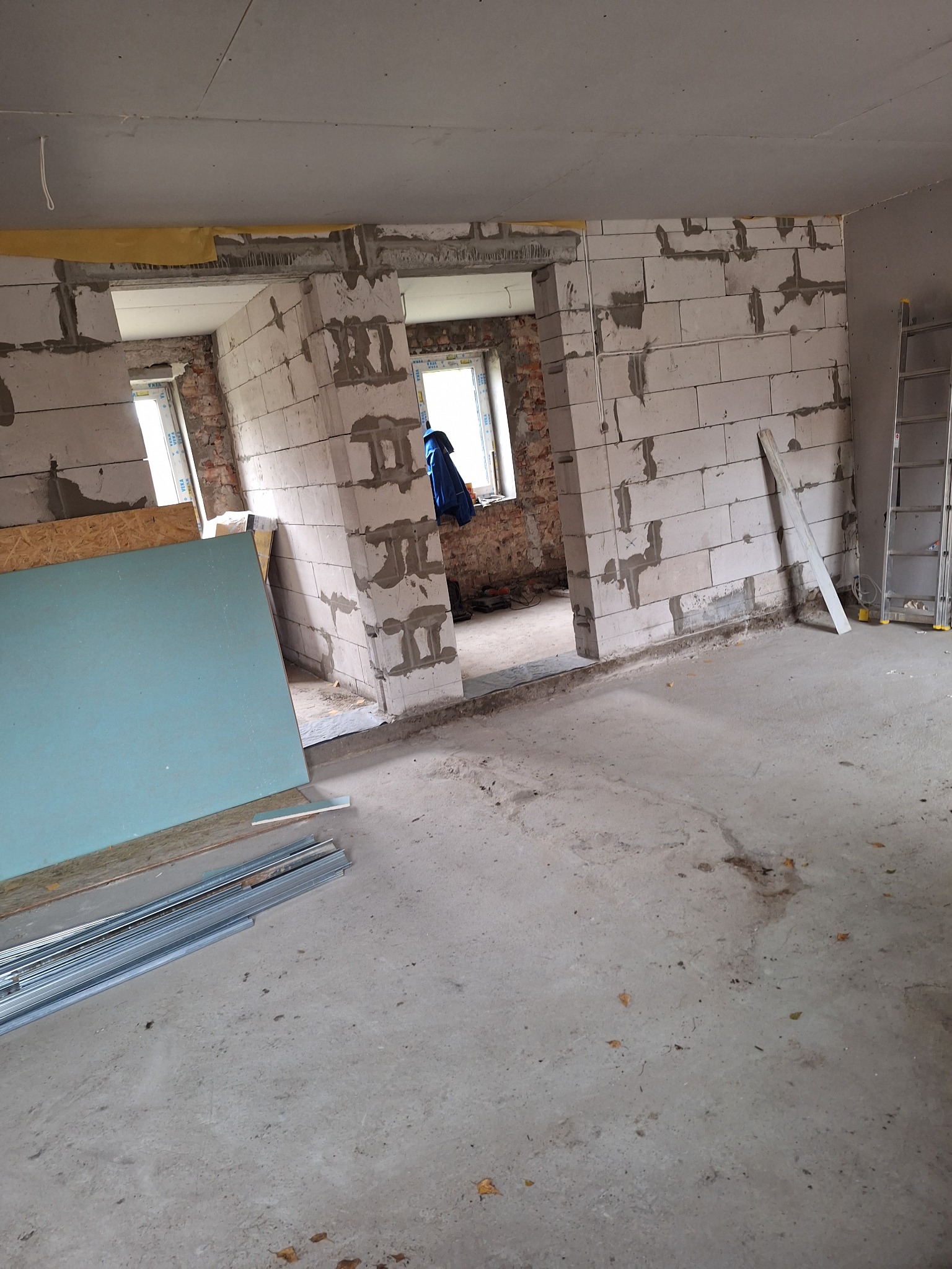 Wnętrze w trakcie remontu z surowymi ścianami z betonu komórkowego, widoczne płyty karton-gips, profile metalowe, oraz drabina oparta o ścianę. W tle okno z widoczną osobą w niebieskiej kurtce.