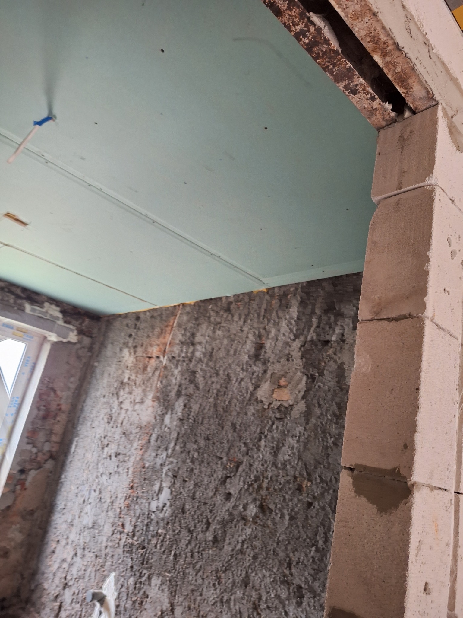 Surowy stan wnętrza w trakcie prac: widoczne płyty na suficie, otynkowana ściana, murowana kolumna i rama drzwiowa z elementami metalowymi.