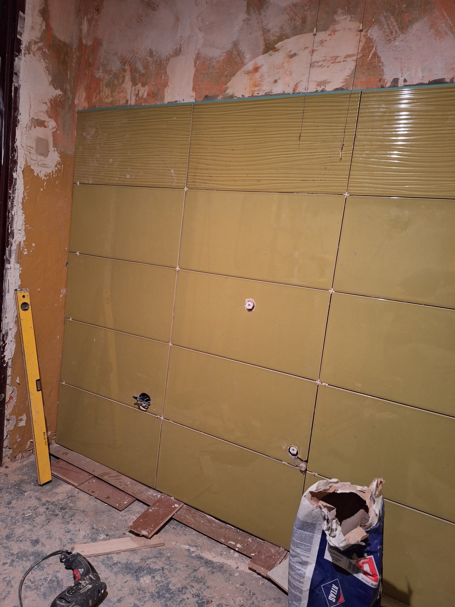 Ściana z oliwkowymi płytkami, częściowo wykończona, z widocznymi otworami na instalacje, poziomica i worek zaprawy na podłodze w trakcie prac w Barwicach.