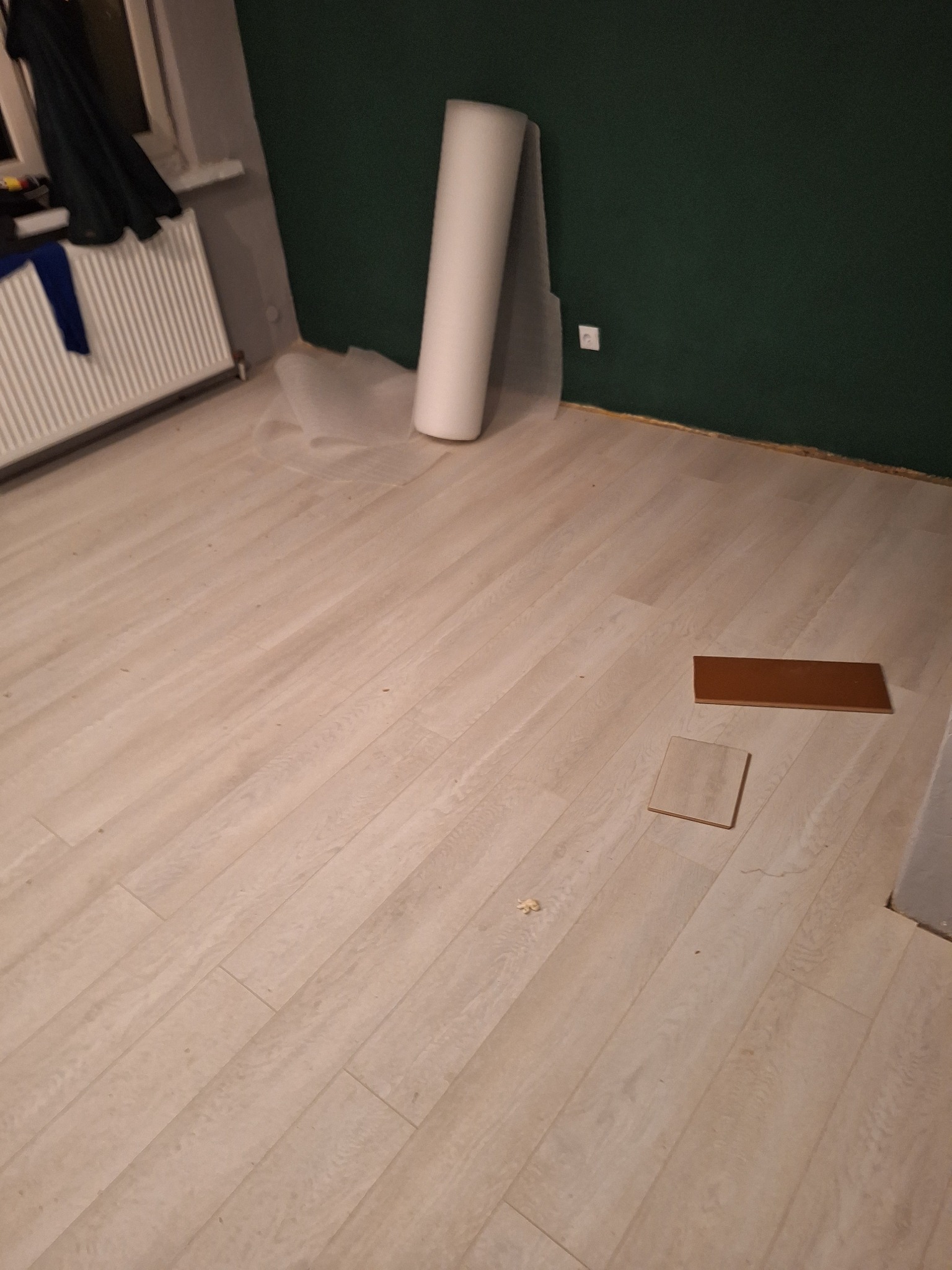 Przygotowanie do położenia jasnych paneli podłogowych, widoczne dwie próbki w odcieniach brązu i rolka folii ochronnej przy ścianie pomalowanej na ciemnozielony kolor.