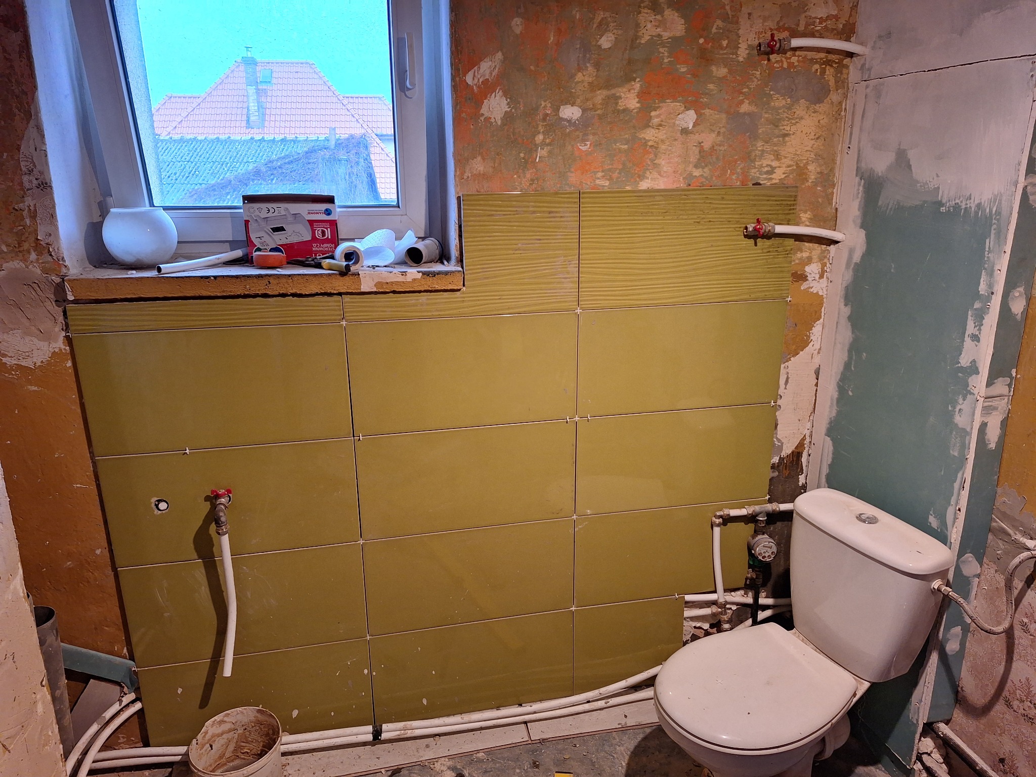 Remont łazienki w trakcie: częściowo ułożone oliwkowe płytki na ścianie, biała toaleta, odsłonięte rury, narzędzia i materiały na parapecie okna z widokiem na dach budynku.