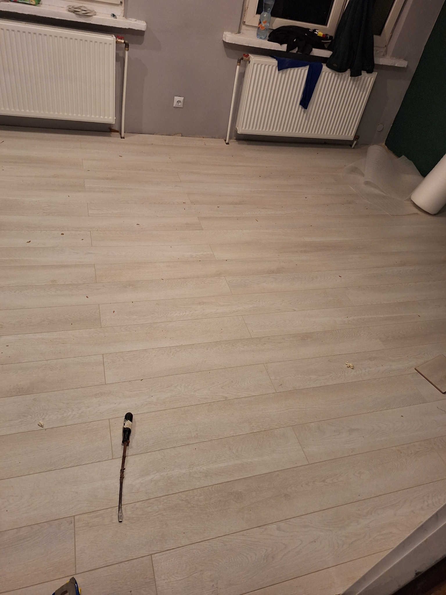 Jasne panele podłogowe ułożone w pokoju z dwoma grzejnikami, widoczny śrubokręt i resztki po montażu.