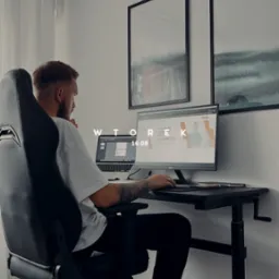 Mężczyzna z tatuażem na ramieniu siedzi na krześle biurowym przed biurkiem z dwoma monitorami, na jednym z nich widoczna strona internetowa z hasłem 'Tworzę strony, które zarabiają'.