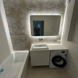 REMOSFERA MYKHAILO HRYN - Wykończona łazienka z wanną, umywalką nablatową, podświetlanym lustrem i pralką. Płytki w odcieniach beżu na ścianie. Widoczne odbicie osoby w lustrze.