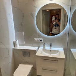 REMOSFERA MYKHAILO HRYN - Wykończona łazienka z marmurowymi płytkami, okrągłym lustrem z podświetleniem LED, białą umywalką z szafką i podwieszaną toaletą. W lustrze odbija się osoba robiąca zdjęcie.