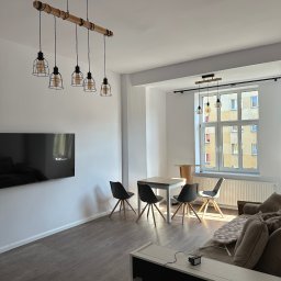 REMOSFERA MYKHAILO HRYN - Jasne wnętrze po remoncie: drewniana lampa wisząca nad stołem z krzesłami, telewizor na ścianie, okno z widokiem na blok. Nowoczesny design, parkiet, neutralna kolorystyka.