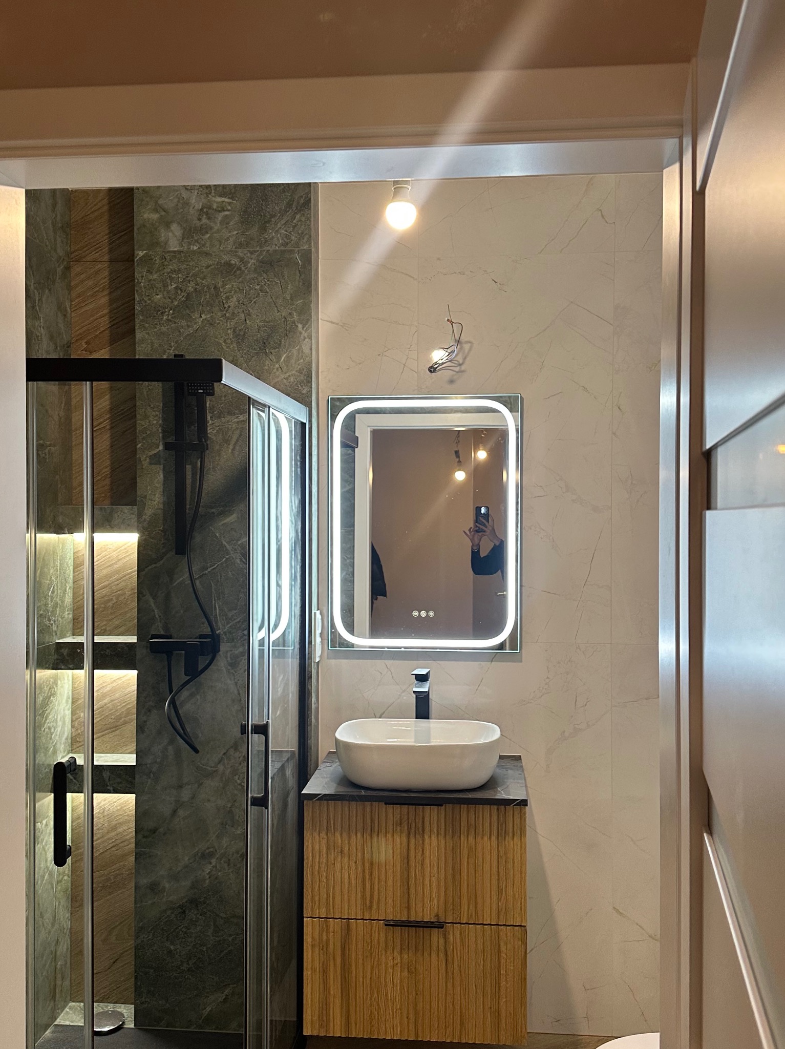 Nowoczesna łazienka po remoncie: kabina prysznicowa z czarnymi elementami, umywalka nablatowa na drewnianej szafce, podświetlane lustro i marmurowe płytki.
