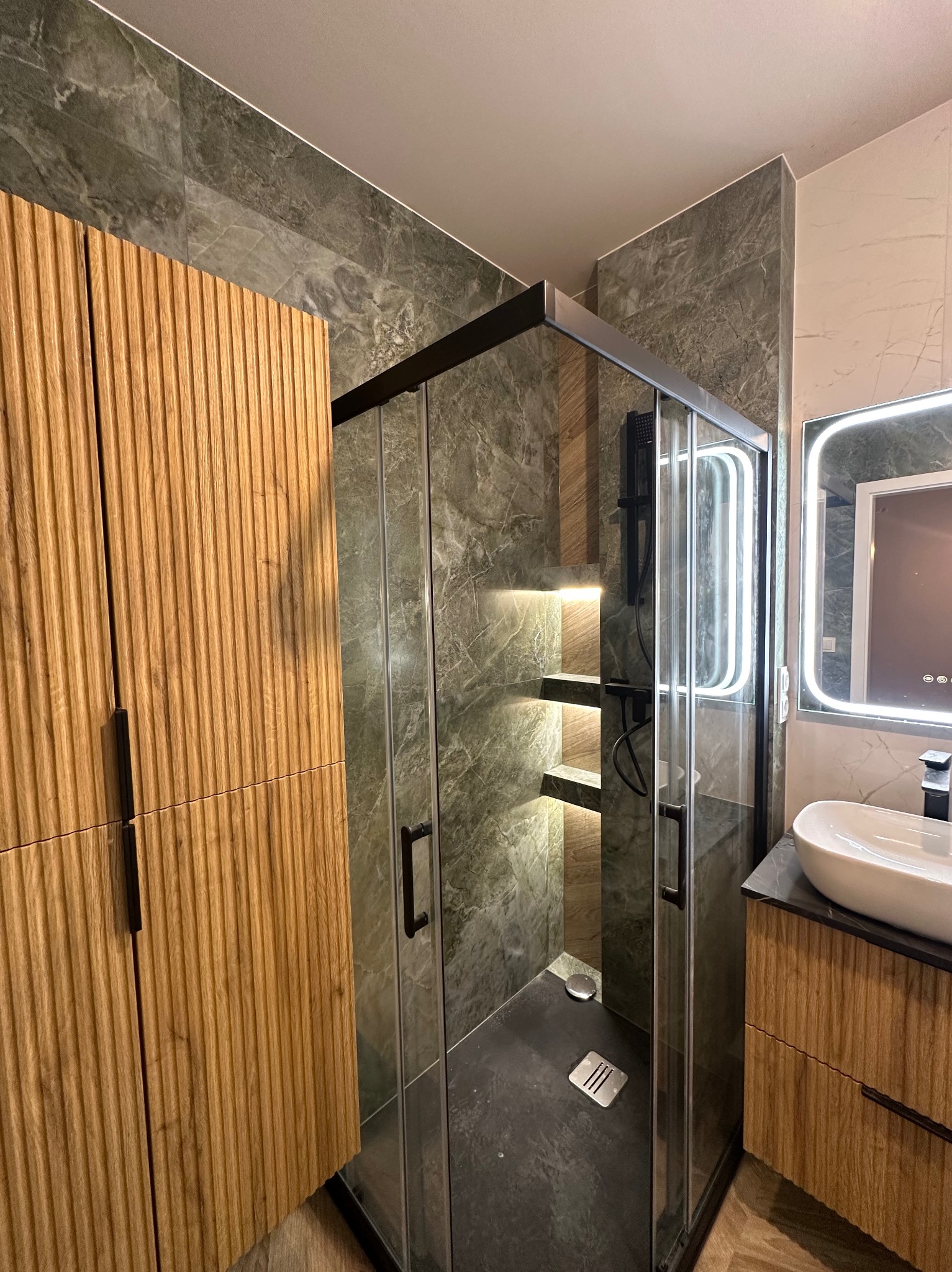 Nowoczesna kabina prysznicowa z czarnymi profilami, marmurowe płytki w odcieniach szarości, drewniana szafka z ryflowanym frontem i podświetlane półki w strefie prysznica.