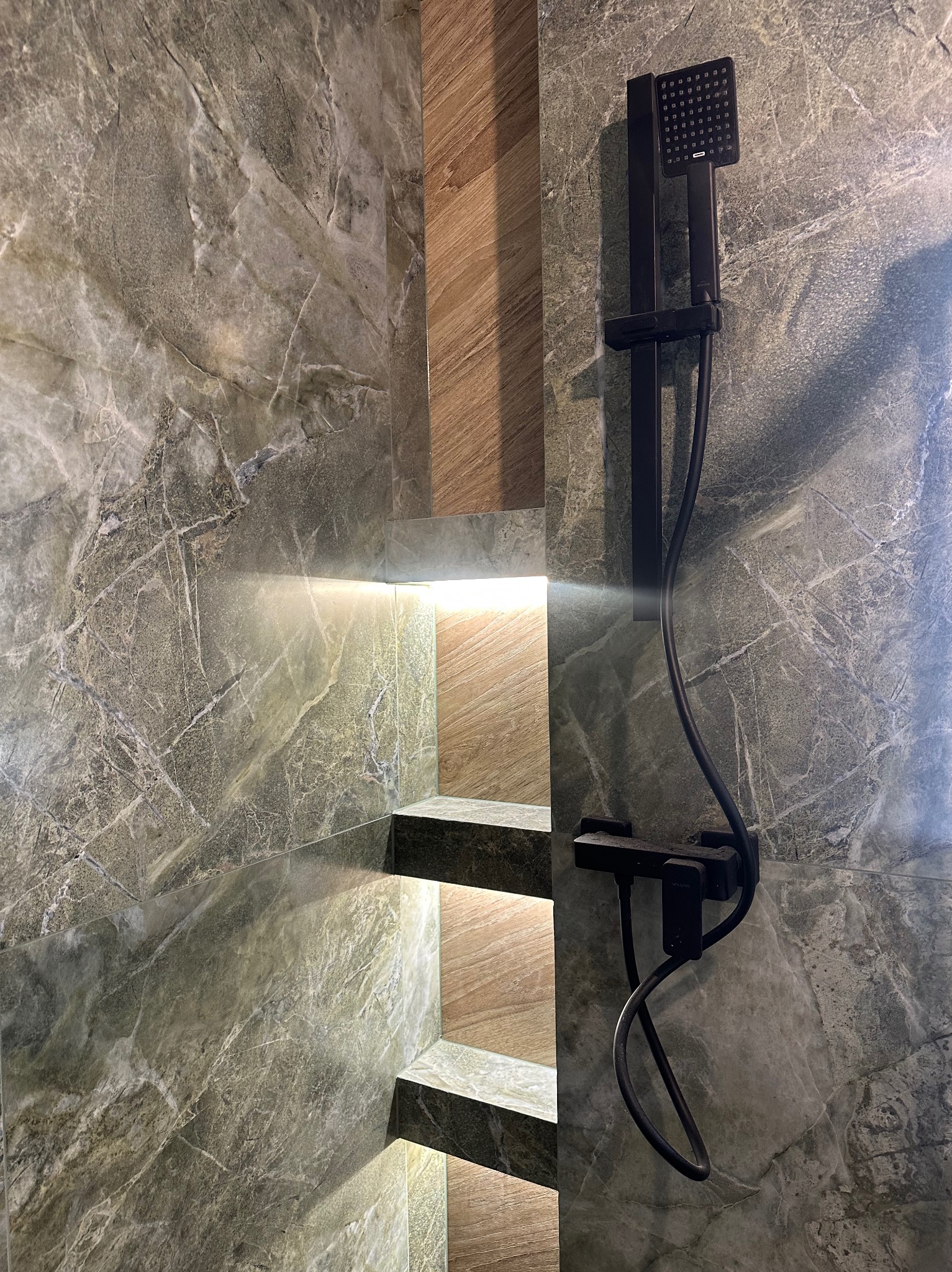 Nowoczesna kabina prysznicowa z szarymi płytkami imitującymi marmur, drewnianymi akcentami i czarną armaturą. Podświetlane półki dodają elegancji.