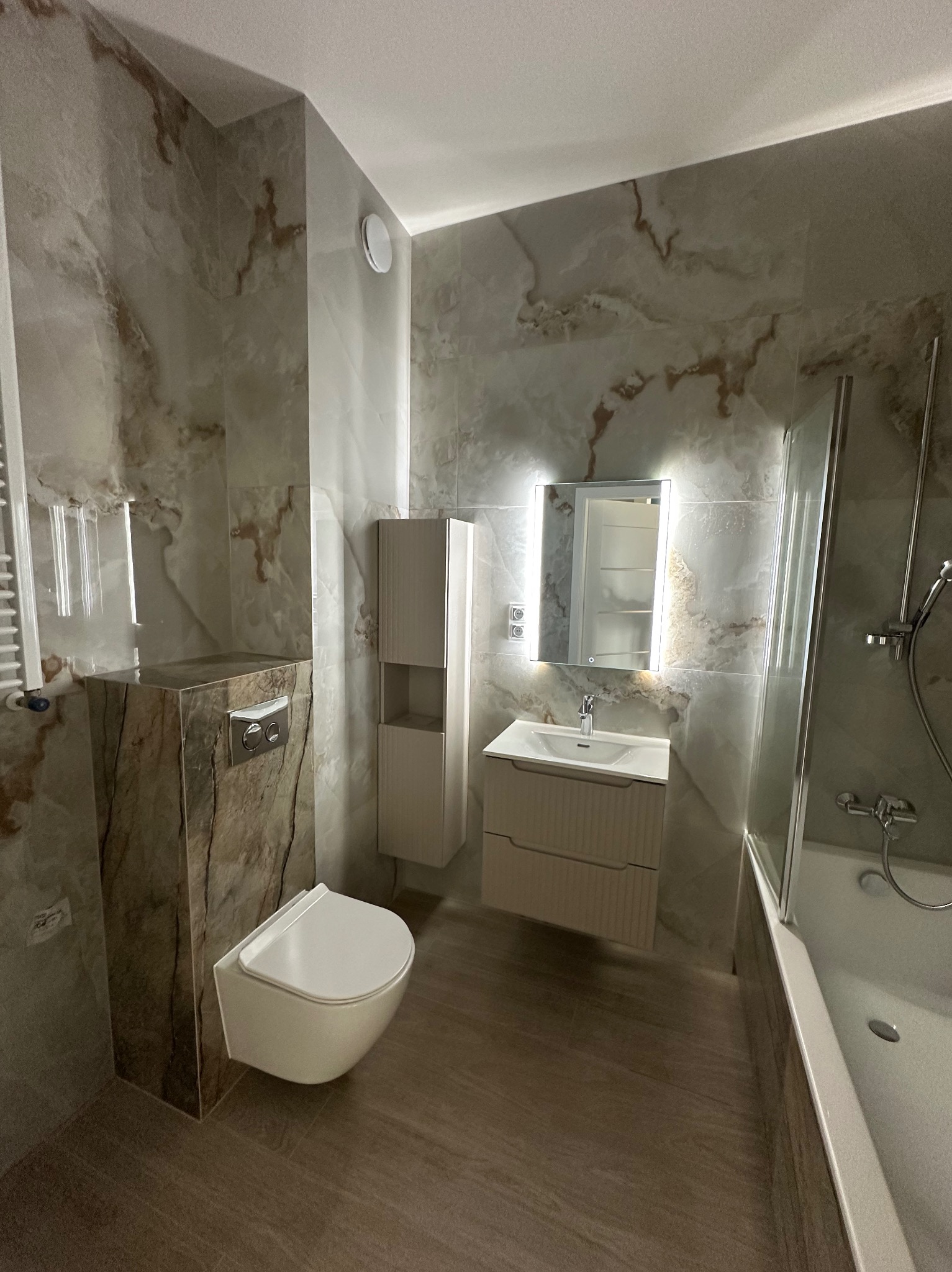Wykończona łazienka z marmurowymi płytkami, podwieszaną toaletą i szafką z lustrem z podświetleniem LED. Nowoczesny design, ciepłe oświetlenie.