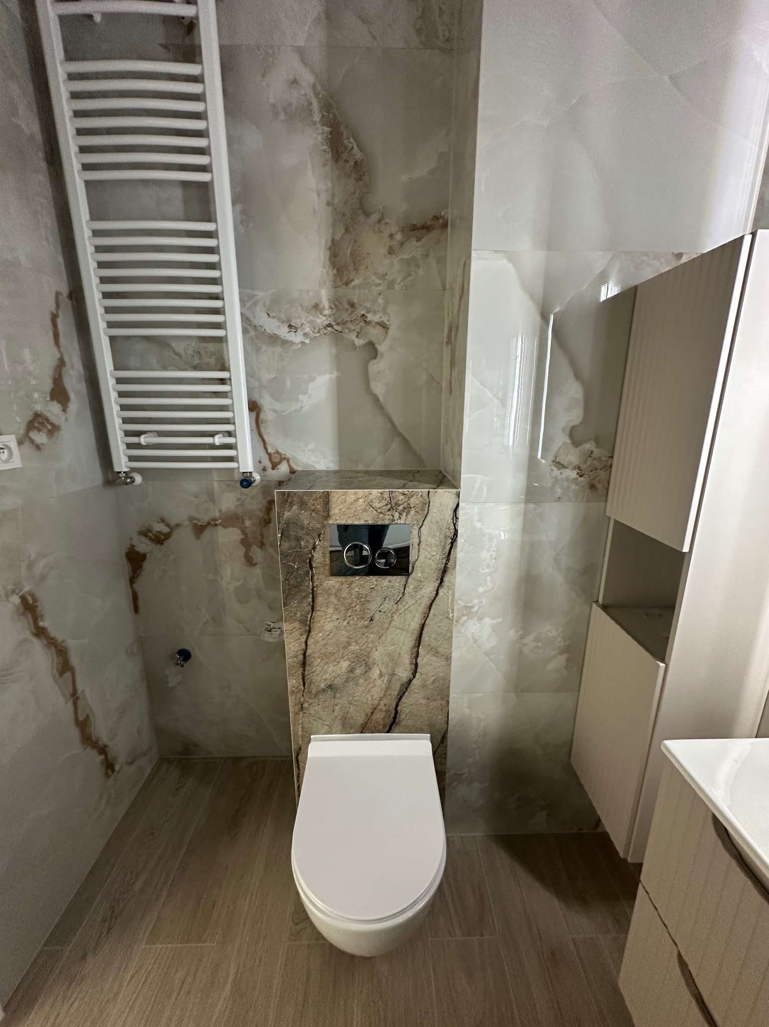 Wykończona łazienka z marmurowymi płytkami, podwieszaną toaletą i grzejnikiem drabinkowym. Umywalka z szafką i szafka wisząca w tle. Nowoczesny design.