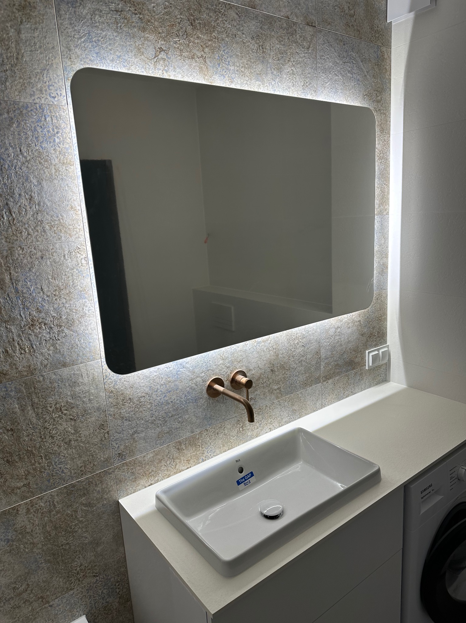 Nowoczesna łazienka we Wrocławiu: umywalka Roca, bateria w kolorze miedzi i podświetlane lustro na tle płytek imitujących kamień.