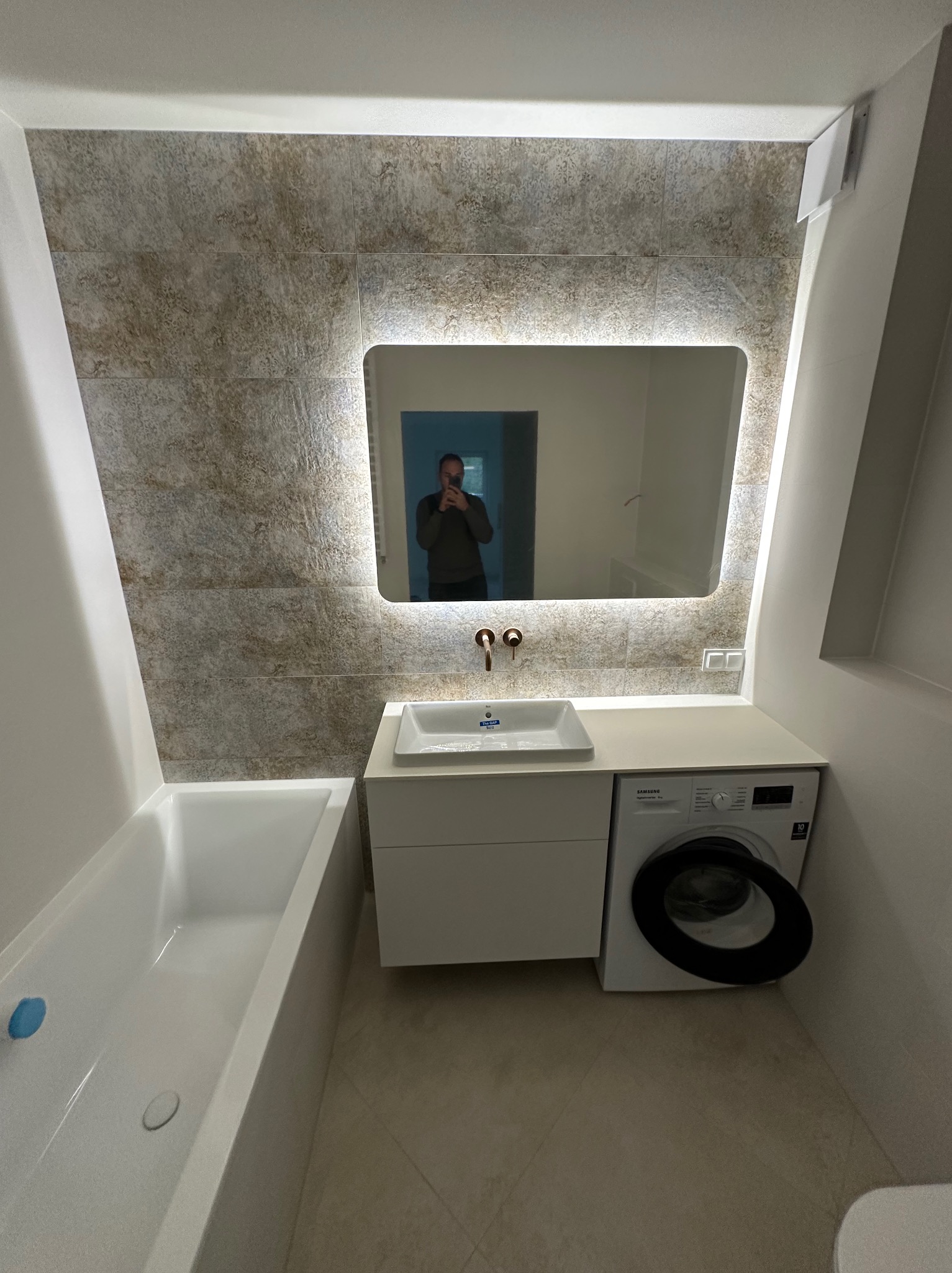 Wykończona łazienka z wanną, umywalką nablatową, podświetlanym lustrem i pralką. Płytki w odcieniach beżu na ścianie. Widoczne odbicie osoby w lustrze.