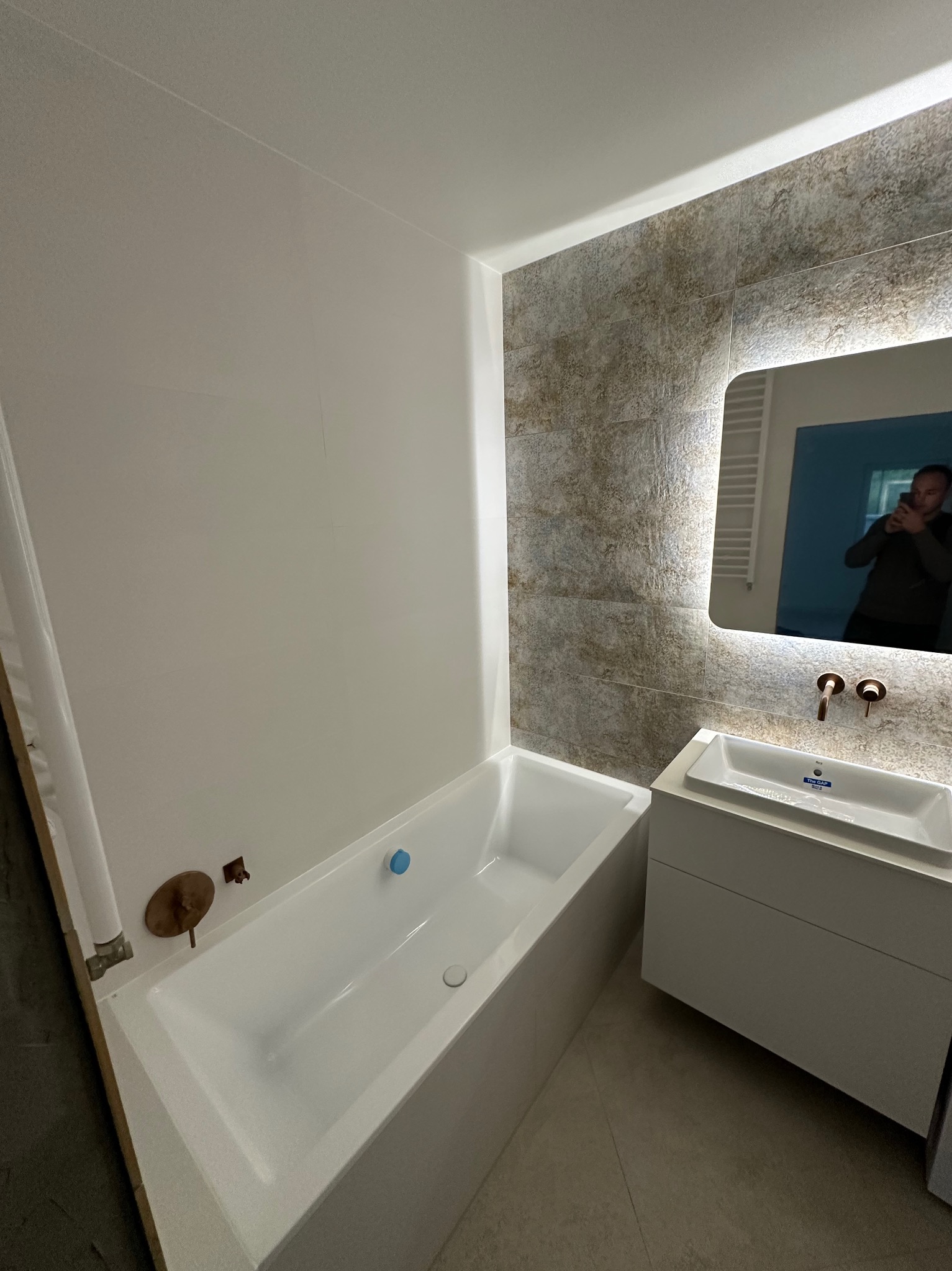 Nowoczesna łazienka z wanną, umywalką na szafce i podświetlanym lustrem. Uwagę zwracają miedziane baterie i płytki imitujące kamień na ścianie.