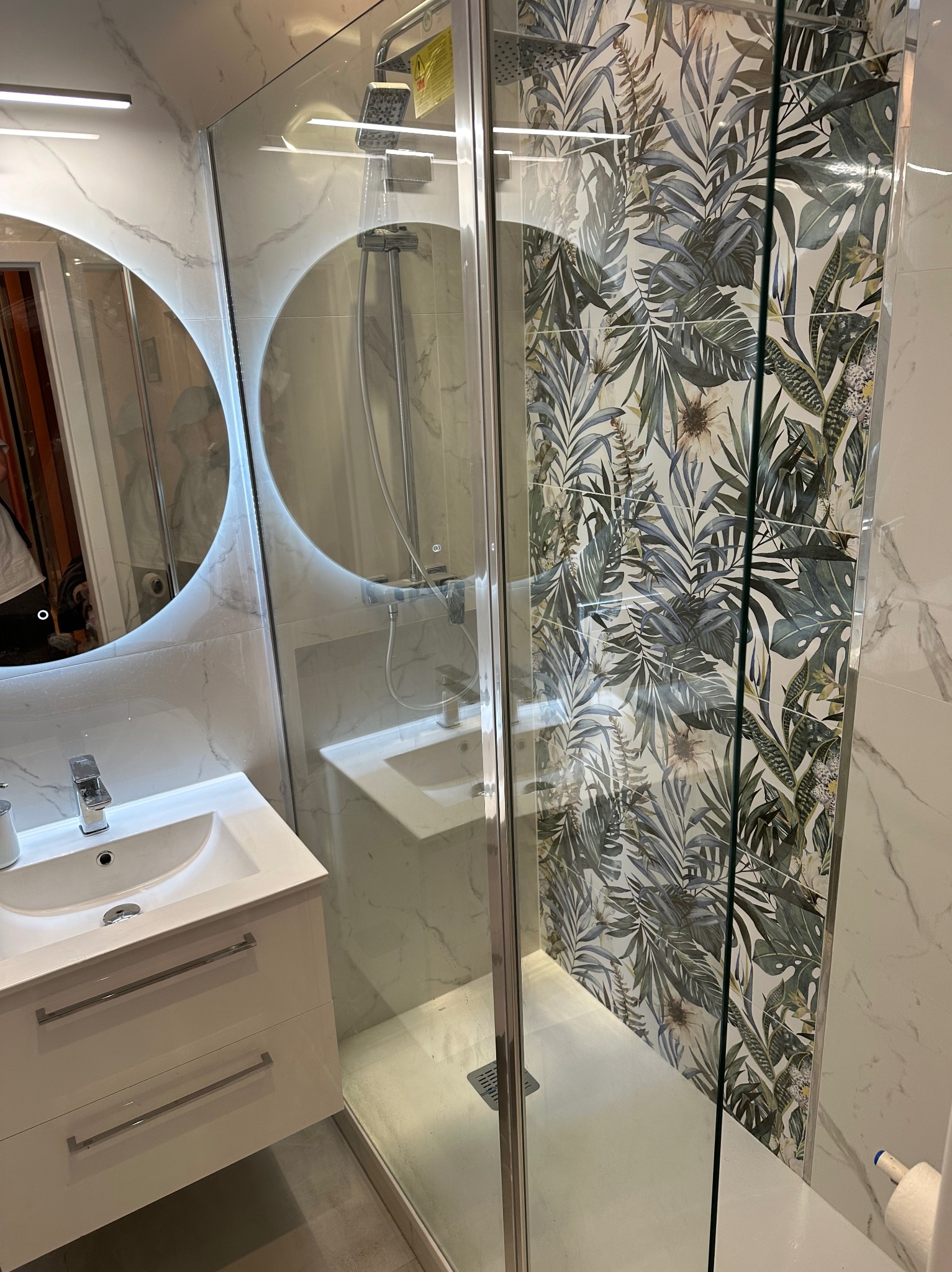 Nowoczesna łazienka po remoncie: kabina prysznicowa ze szklaną ścianą i płytkami w tropikalne liście, okrągłe lustro z podświetleniem LED nad białą szafką z umywalką.