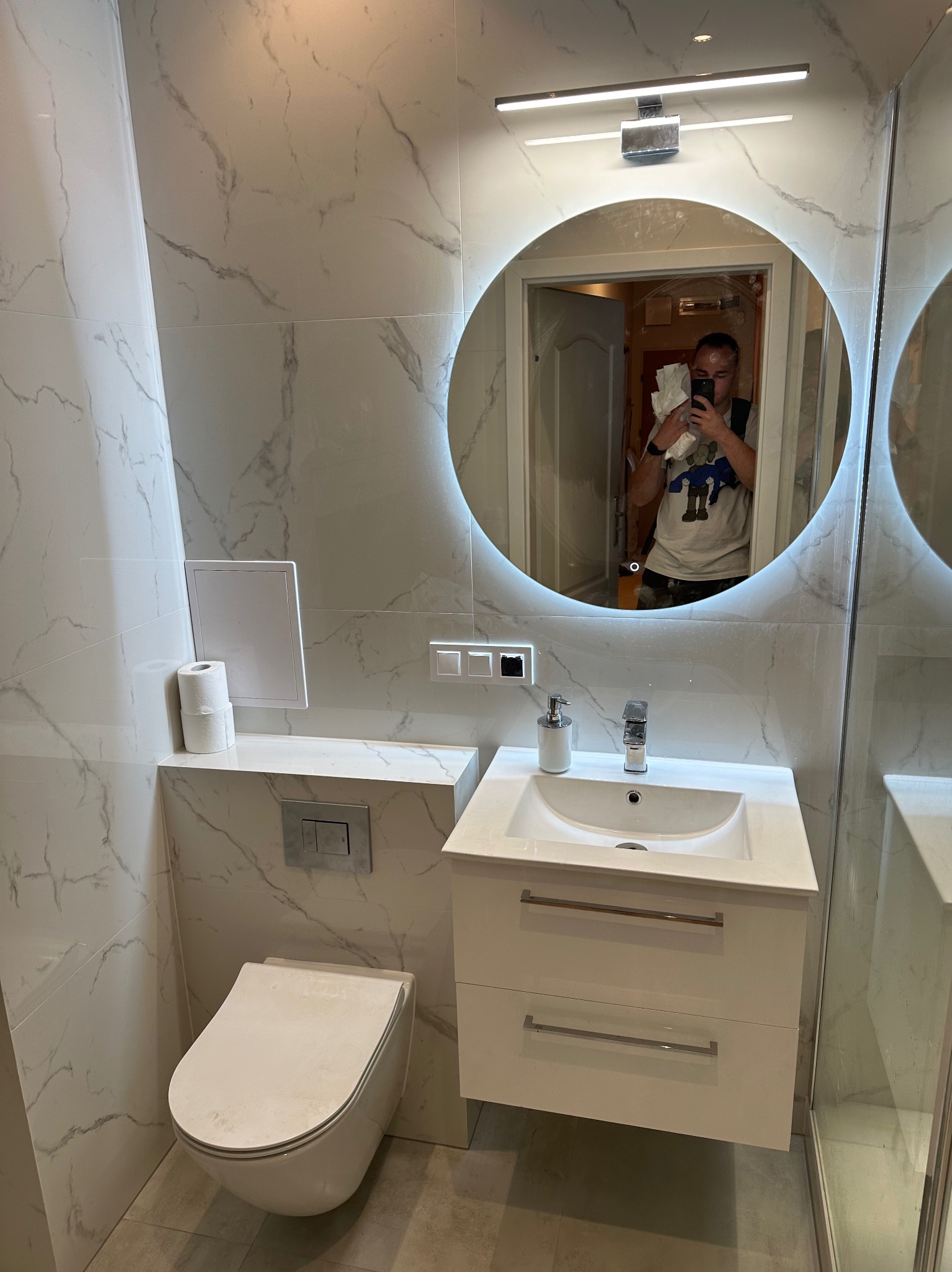 Wykończona łazienka z marmurowymi płytkami, okrągłym lustrem z podświetleniem LED, białą umywalką z szafką i podwieszaną toaletą. W lustrze odbija się osoba robiąca zdjęcie.