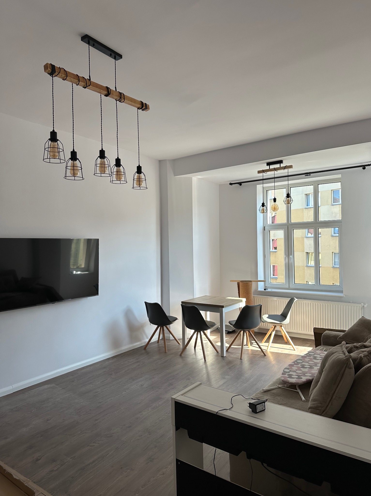 Jasne wnętrze po remoncie: drewniana lampa wisząca nad stołem z krzesłami, telewizor na ścianie, okno z widokiem na blok. Nowoczesny design, parkiet, neutralna kolorystyka.