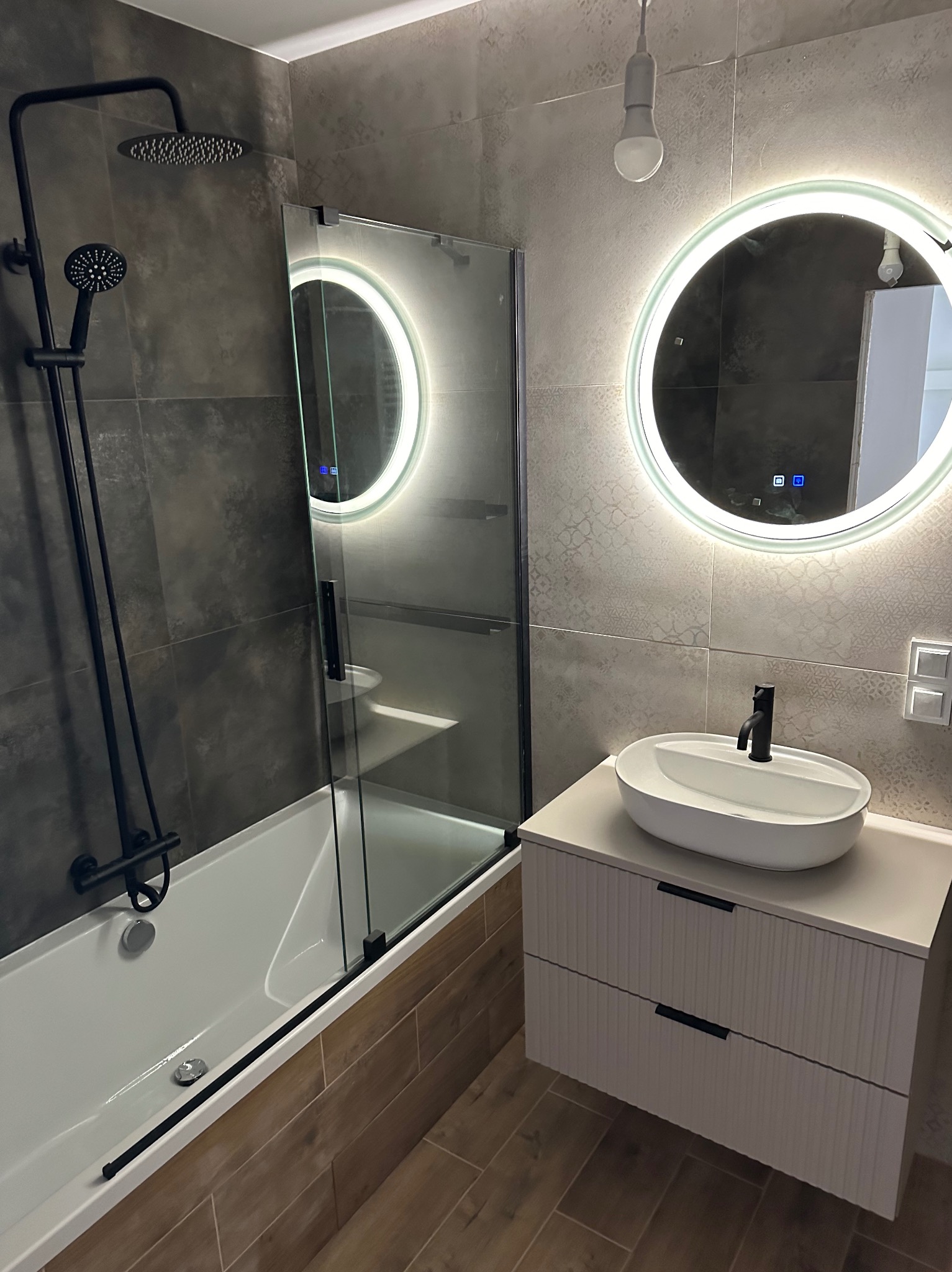 Nowoczesna łazienka z wanną, szklanym parawanem, umywalką na szafce z frezowanymi frontami i okrągłym lustrem z podświetleniem LED. Ciemne płytki na ścianach.