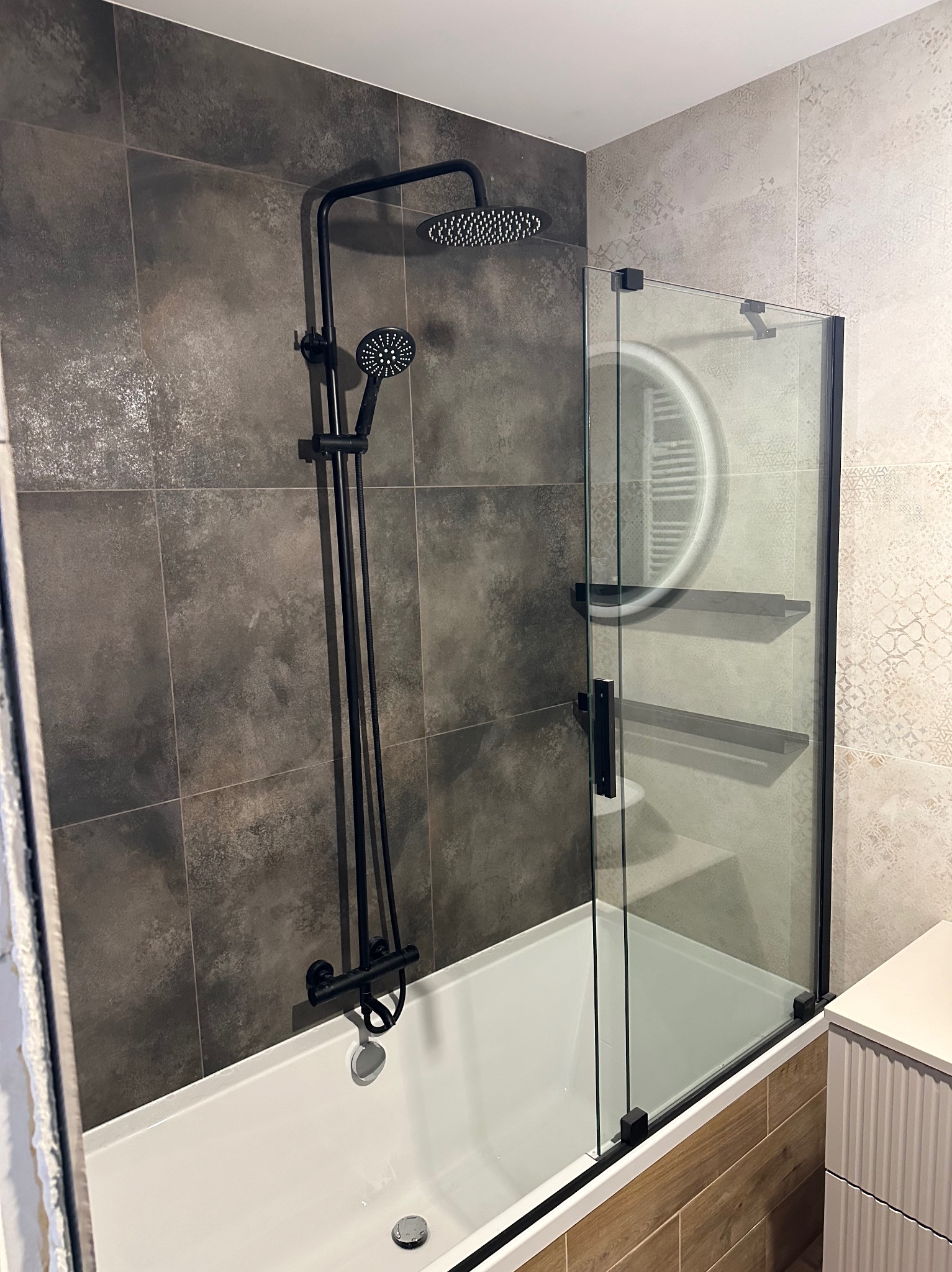 Nowoczesna łazienka po remoncie: biała wanna z czarną armaturą i szklanym parawanem, płytki w odcieniach szarości i beżu, oświetlenie LED. Minimalistyczny design.