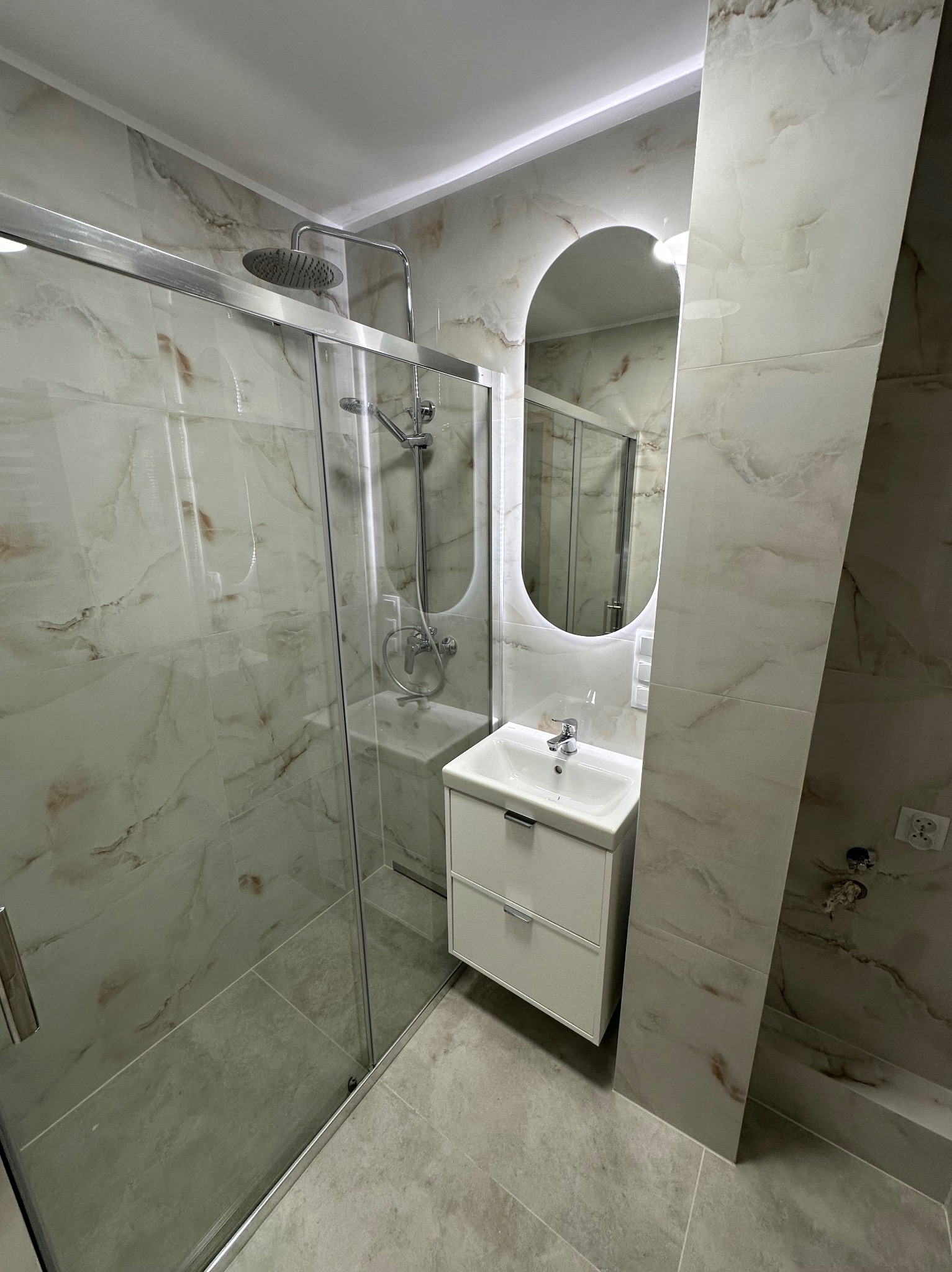 Nowoczesna łazienka po remoncie: kabina prysznicowa ze szklanymi drzwiami, umywalka z szafką i podświetlane lustro na ścianie wyłożonej marmurkowymi płytkami.