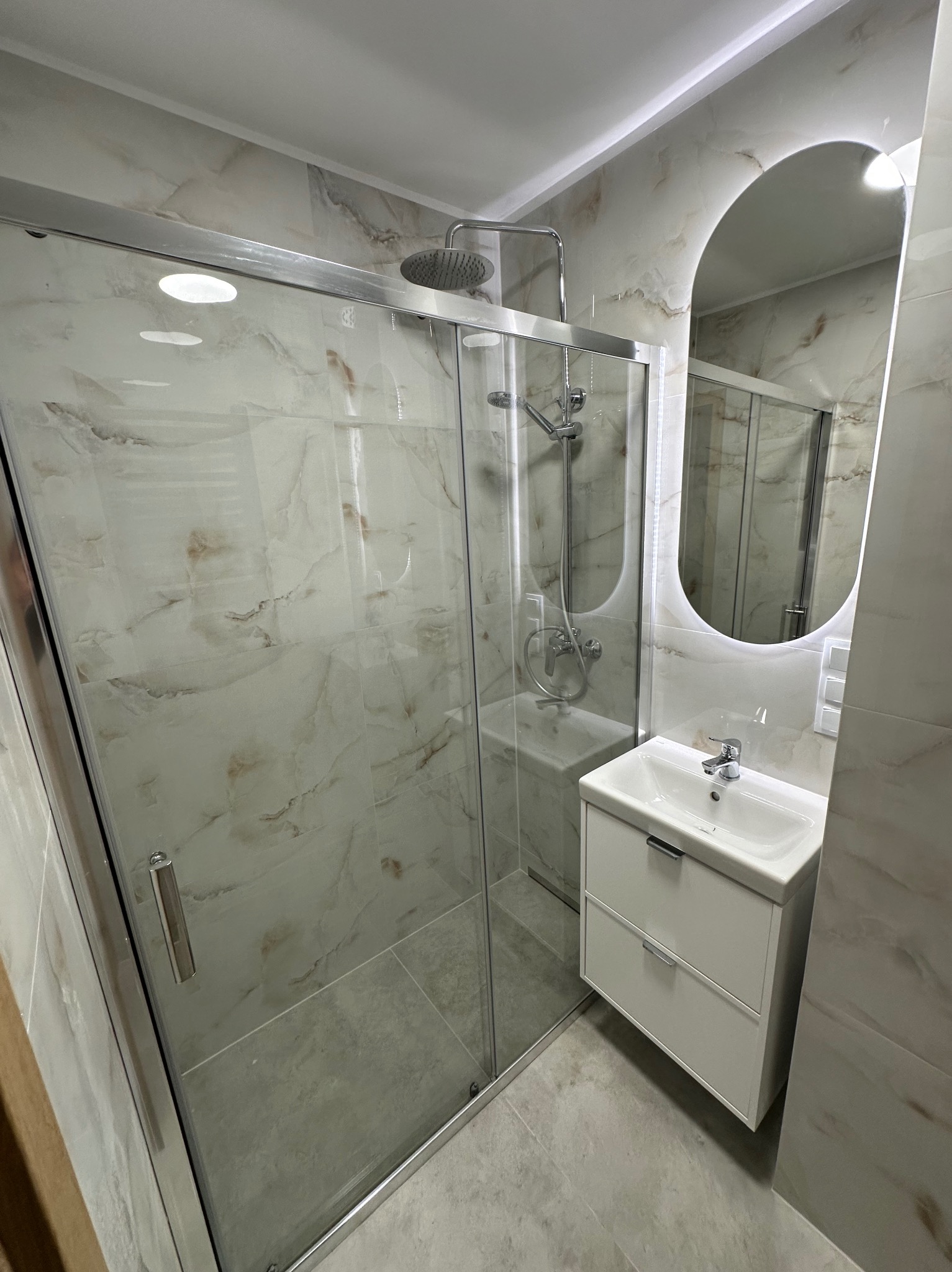 Nowoczesna łazienka z kabiną prysznicową typu walk-in, umywalką z szafką i podświetlanym lustrem. Marmurowe płytki na ścianach i podłodze. Minimalistyczny design.