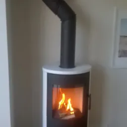 Nowoczesny kominek wolnostojący z czarnym korpusem i białym obramowaniem, z widocznym płomieniem, zainstalowany w domu w Wolicy, z drewnianą podłogą i szklaną płytą pod kominkiem.