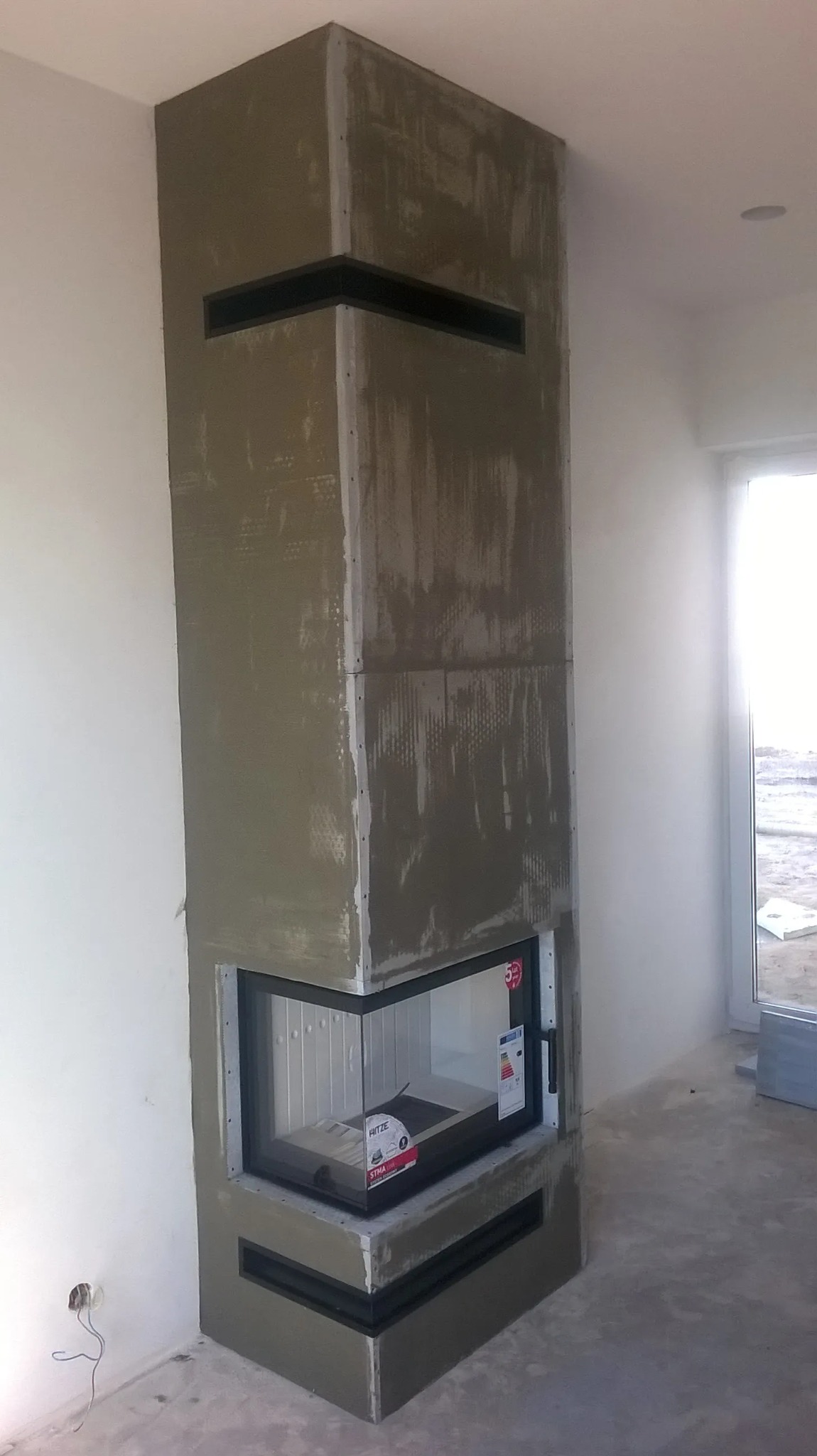 Kominek w trakcie zabudowy, widoczne płyty i warstwa kleju, z czarnymi poziomymi wstawkami wentylacyjnymi, z szybą i wkładem kominkowym w środku, kabel elektryczny wystaje ze ściany.