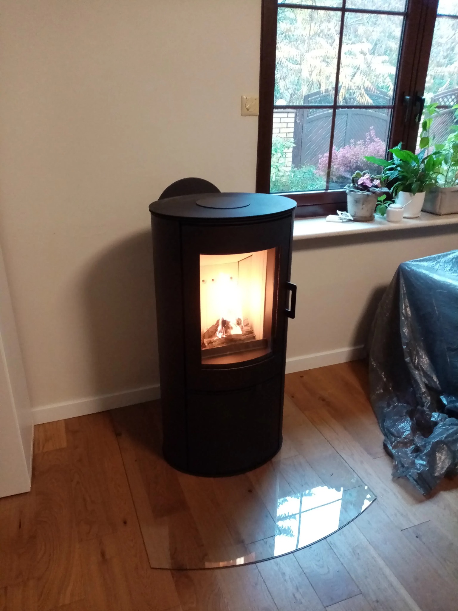 Czarny, cylindryczny kominek wolnostojący z palącym się drewnem, na przezroczystej półokrągłej płycie ochronnej na drewnianej podłodze, z widokiem przez okno na zewnątrz