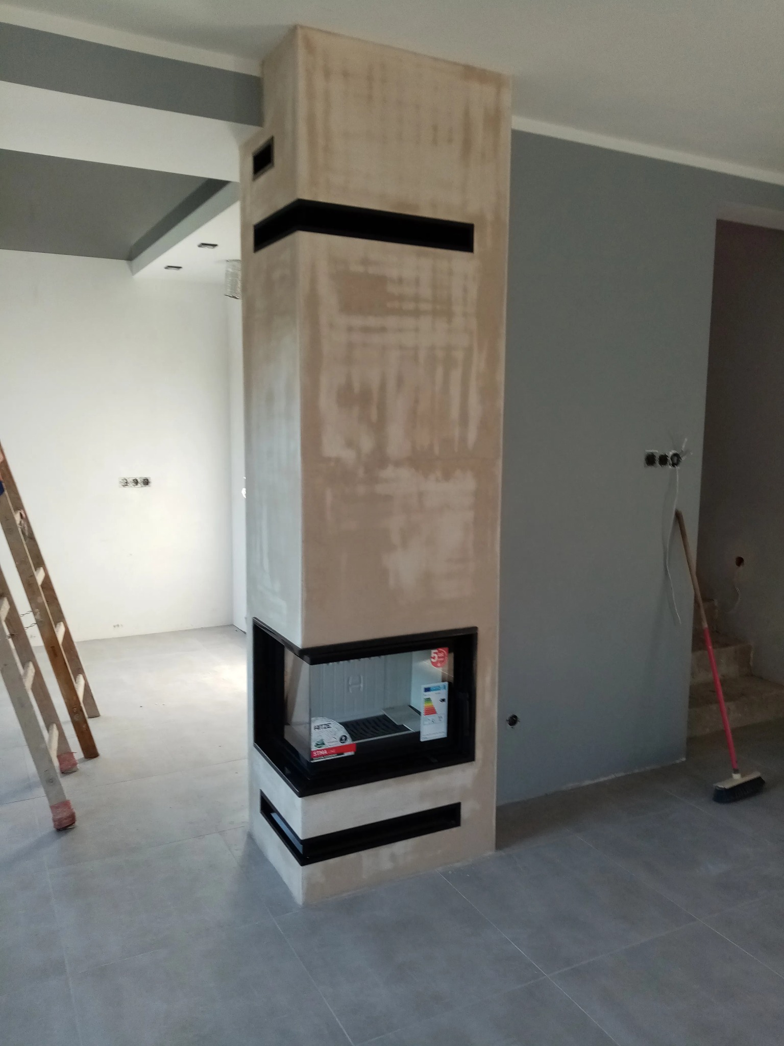 Nowoczesny kominek narożny z czarnymi akcentami w surowym wykończeniu, widoczna drabina malarska i niedokończone ściany w szaro-białej kolorystyce.