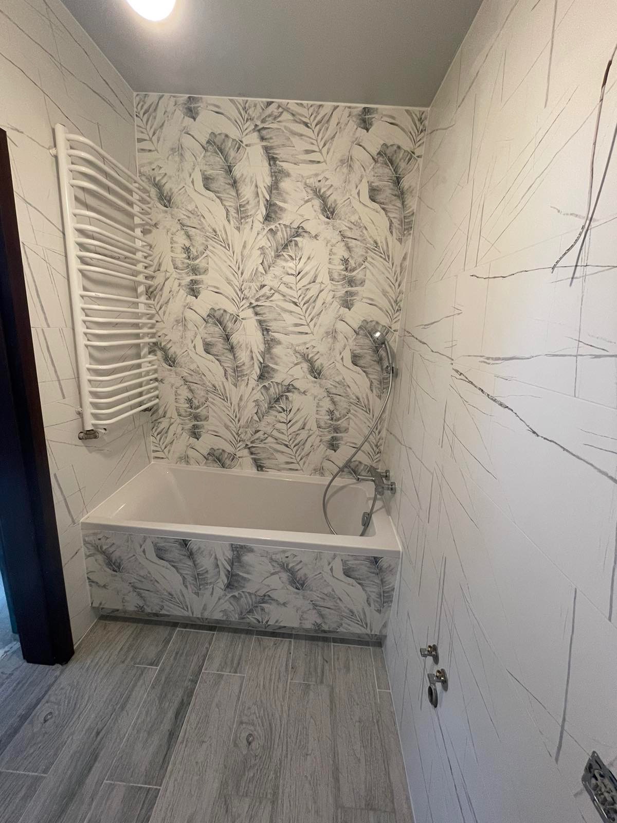 Wykończona łazienka z wanną obudowaną płytkami w motywie liści, grzejnikiem drabinkowym i szarymi panelami podłogowymi imitującymi drewno.