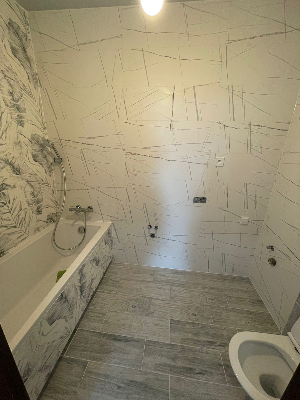 Wykończona łazienka z białymi płytkami imitującymi marmur z szarymi żyłkami, oraz płytkami podłogowymi imitującymi drewno w odcieniach szarości. Wanna obudowana płytkami z motywem liści. Widoczne...